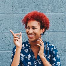 Woman with bright red hair smiles, pointing towards the left against a blue brick wall.