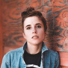 Person with short brown hair wearing a denim jacket, leaning against a weathered orange wall.