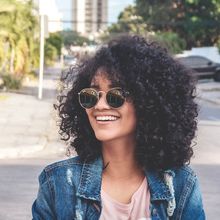 Woman with curly hair and sunglasses smiles, wearing a denim jacket, outdoors on a city street.