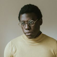 Man wearing round gold-rimmed glasses and a yellow turtleneck looks off-camera with a thoughtful expression.
