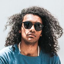 Man with curly hair and sunglasses wearing a blue shirt and a beaded necklace.