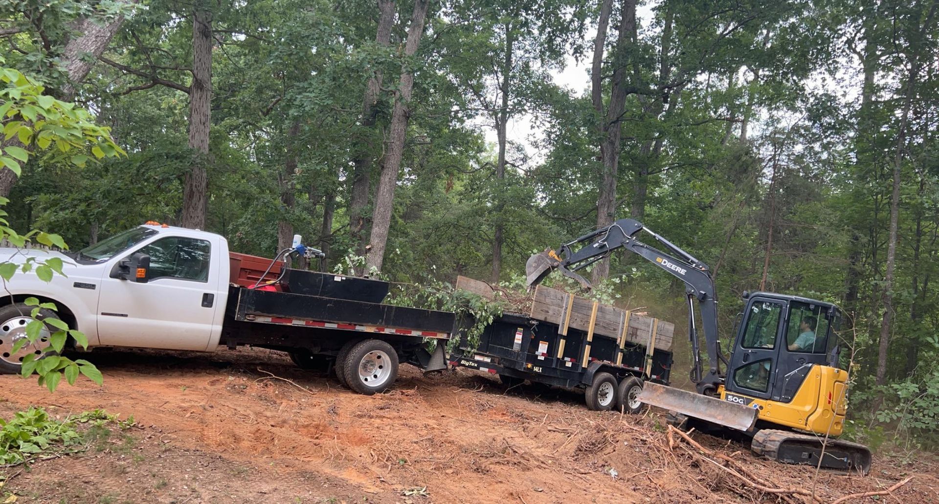 A white flatbed truck towing a trailer as a yellow excavator loads debris into it on a dirt forest trail.