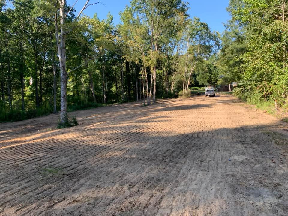 A recently cleared, level dirt lot with tire tracks, surrounded by lush green trees under a bright blue sky.