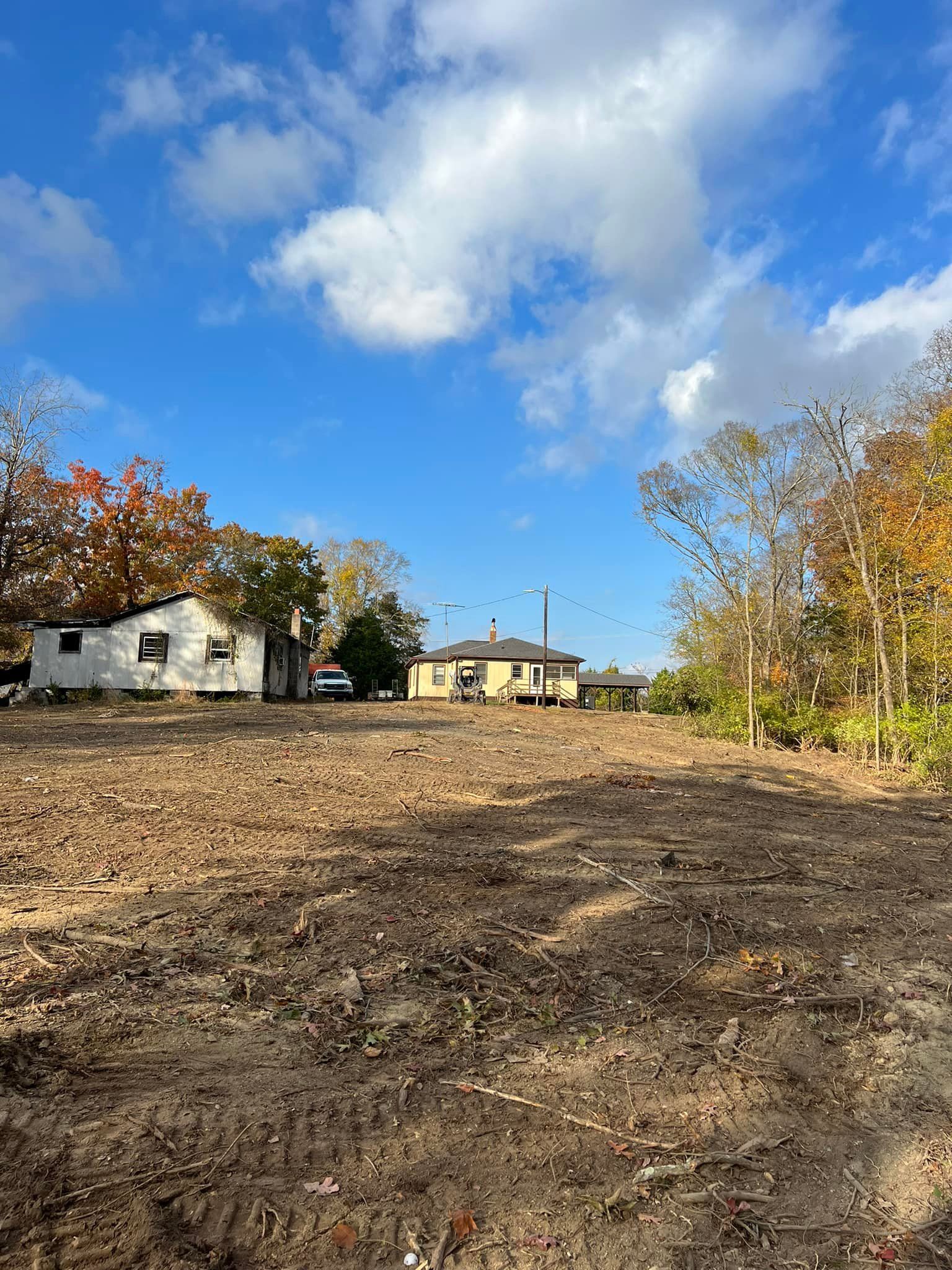 A cleared, dark soil lot in the foreground, with two small houses visible in the background under a sunny, cloudy sky.