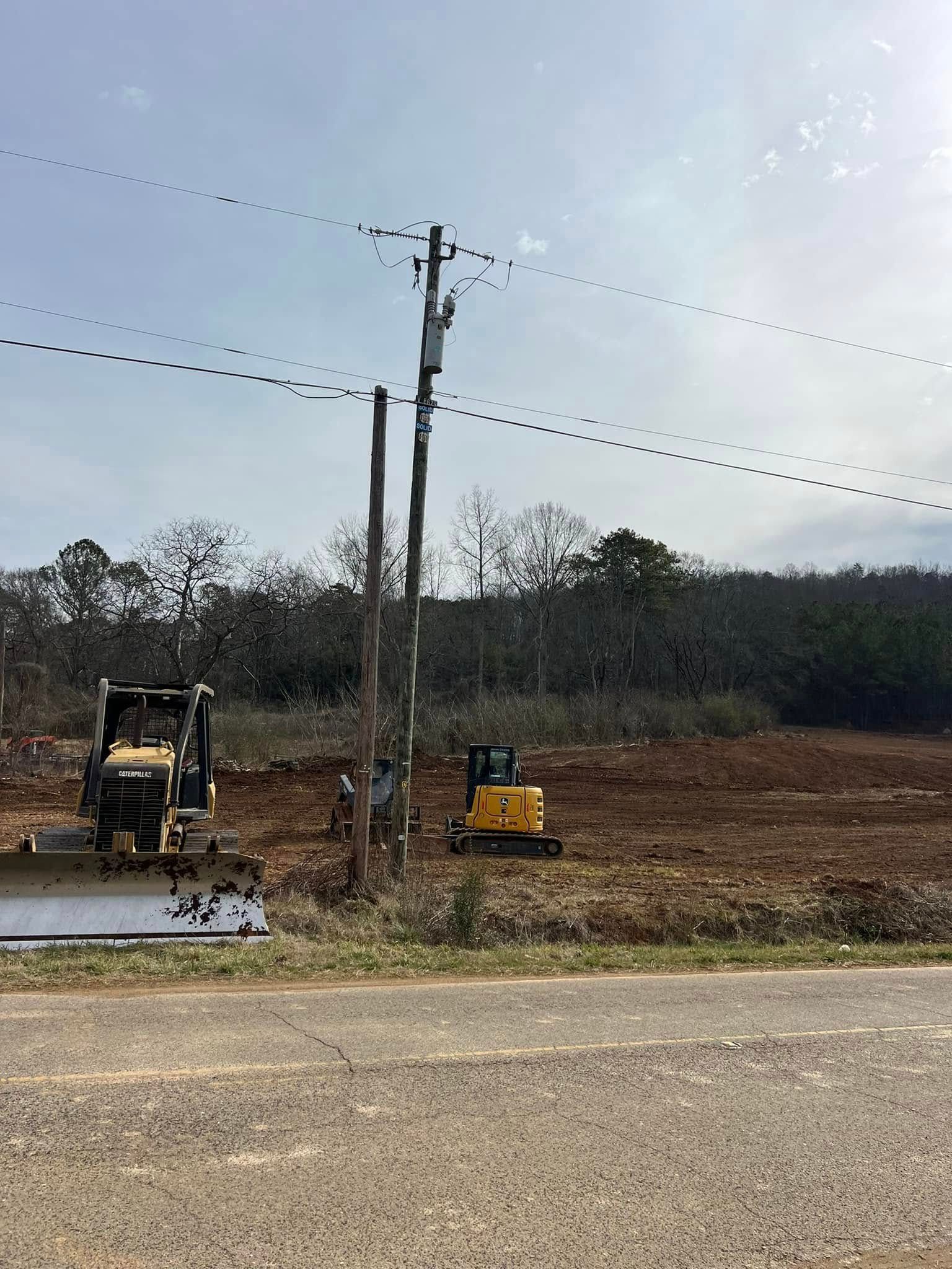 Two yellow construction vehicles work in a clearing next to a utility pole and a gravel road under a bright sky.
