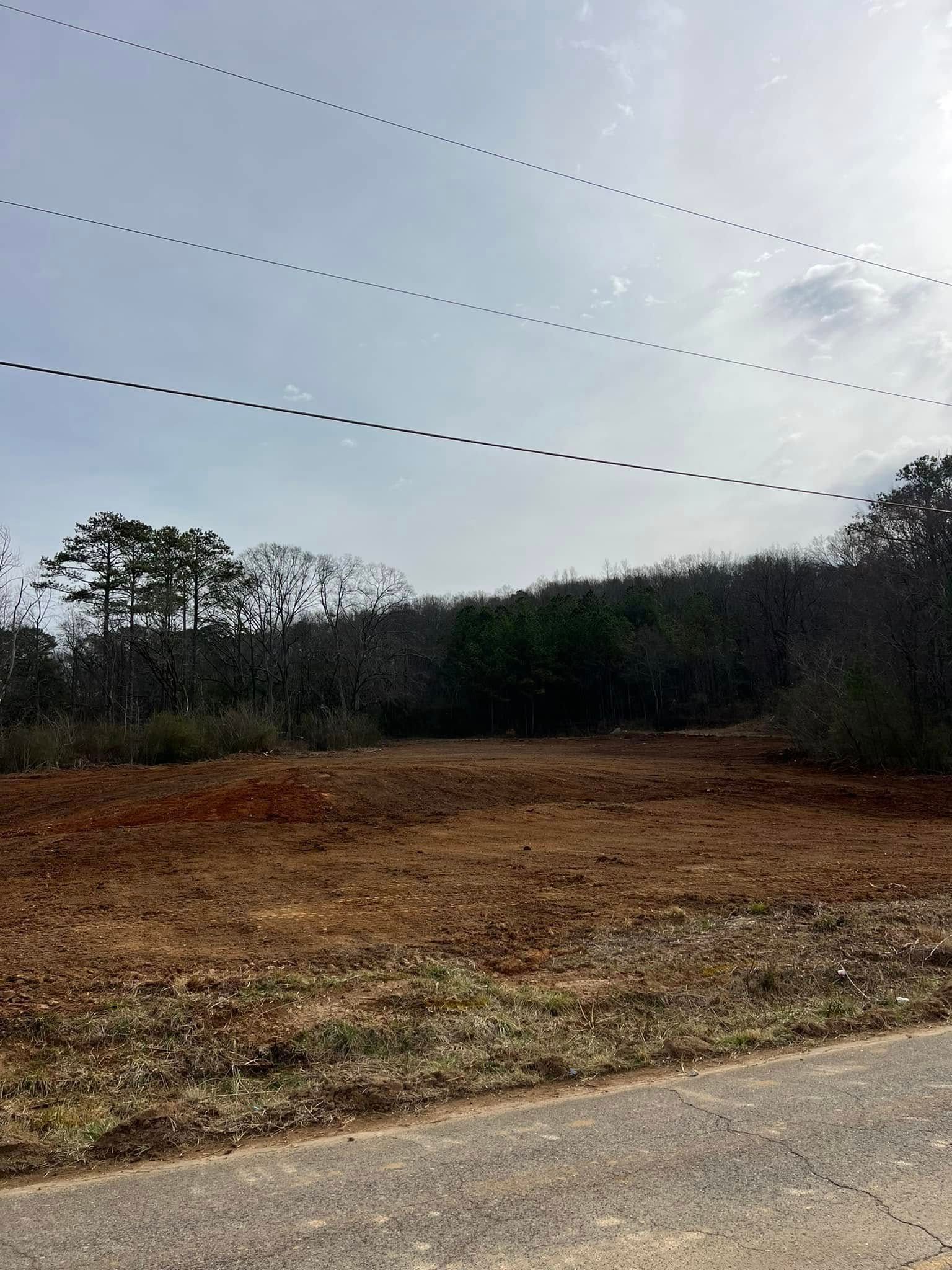 A clearing of bare red-brown soil bordered by trees under a bright, cloudy sky with overhead power lines.