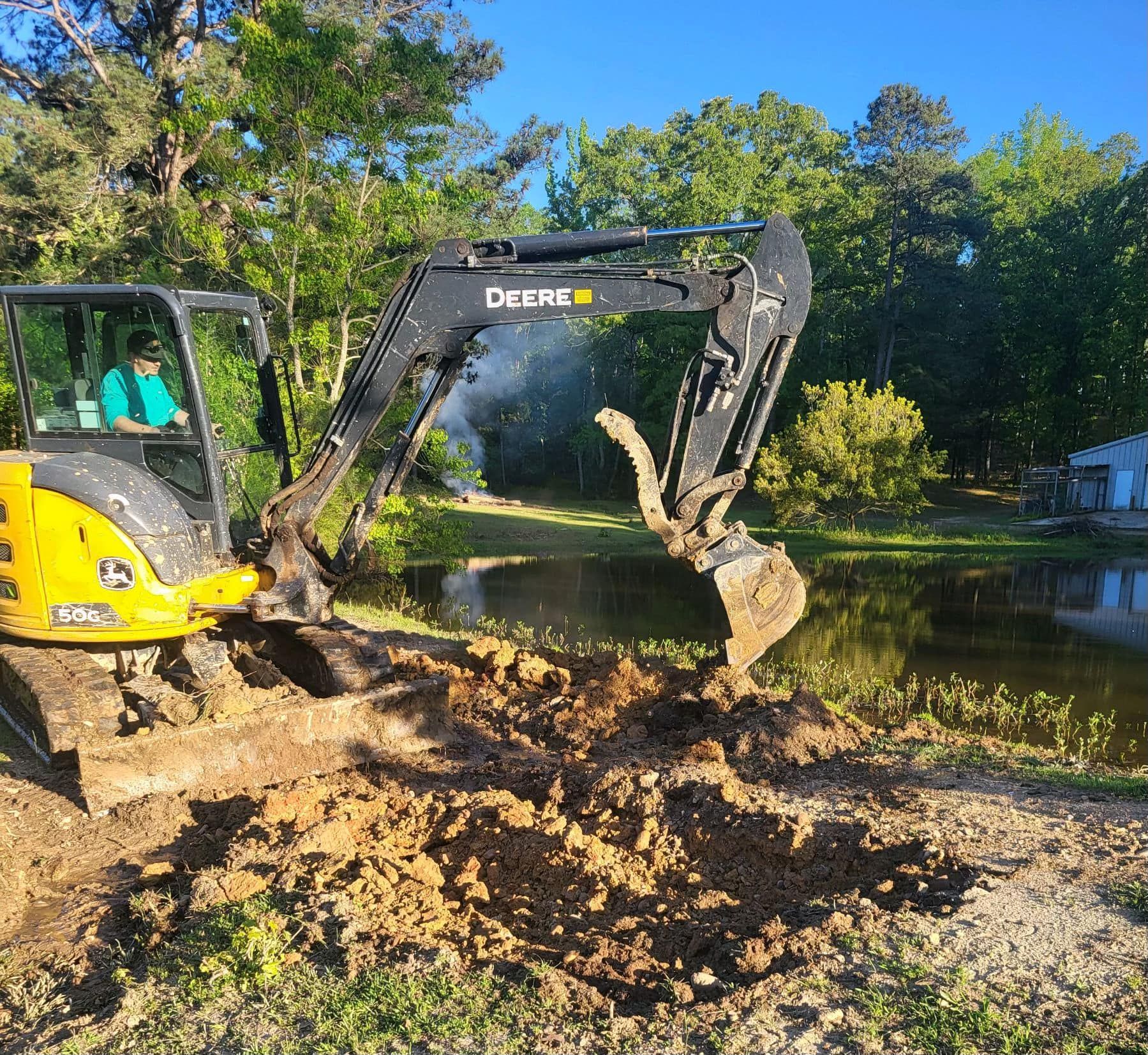 A yellow John Deere excavator digs into the muddy bank of a pond on a sunny day with trees in the background.