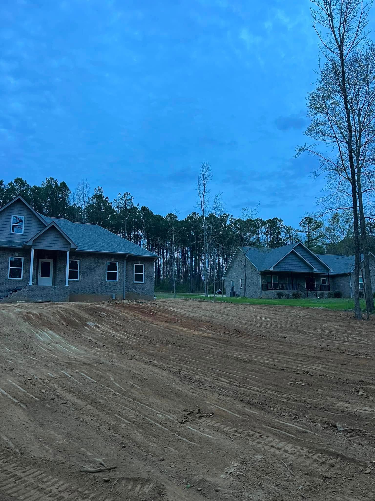 Two new gray brick houses stand under a twilight sky, separated by a vast, bare field of dirt and gravel.