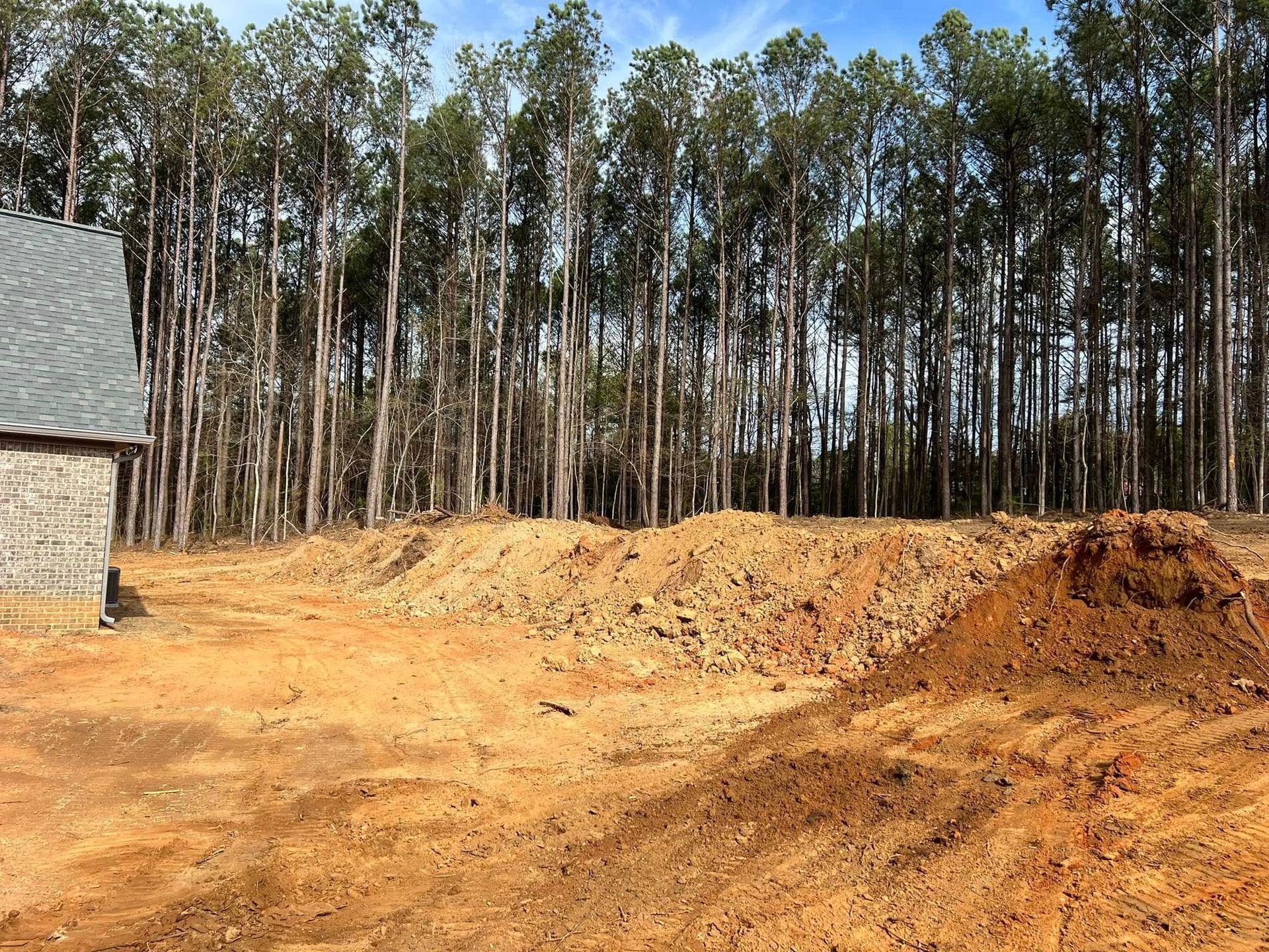 A residential construction site with large piles of dirt in the foreground and a dense forest in the background.