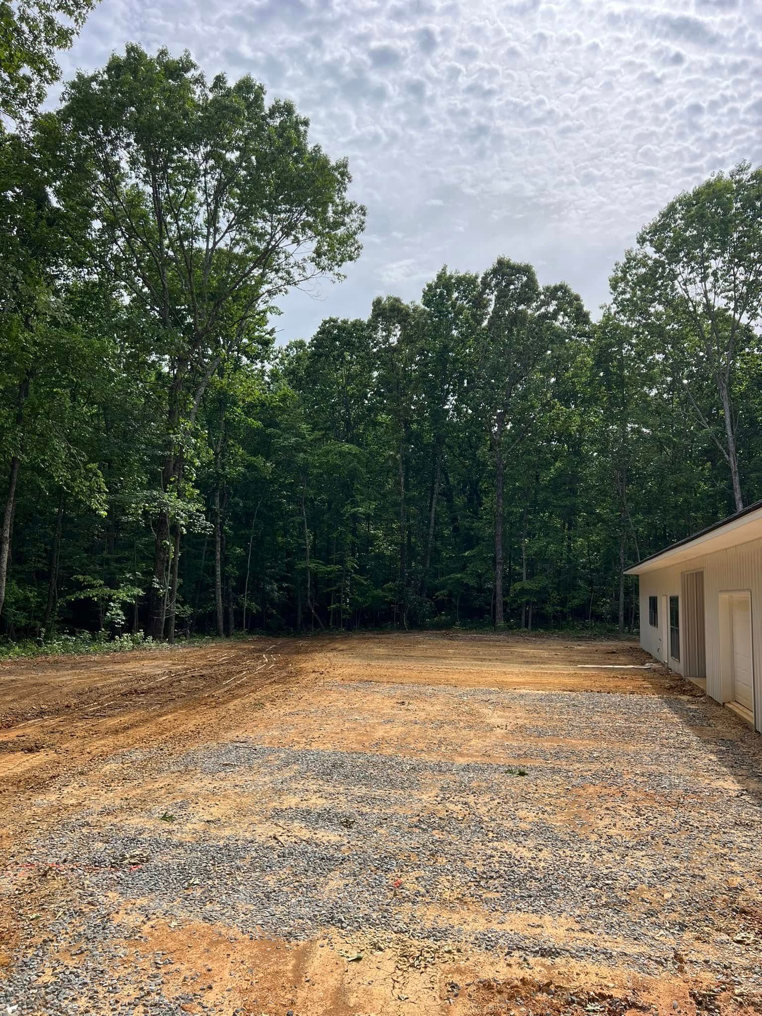 A gravel-filled area sits in front of a line of tall, green trees next to the side of a white building under a cloudy sky.