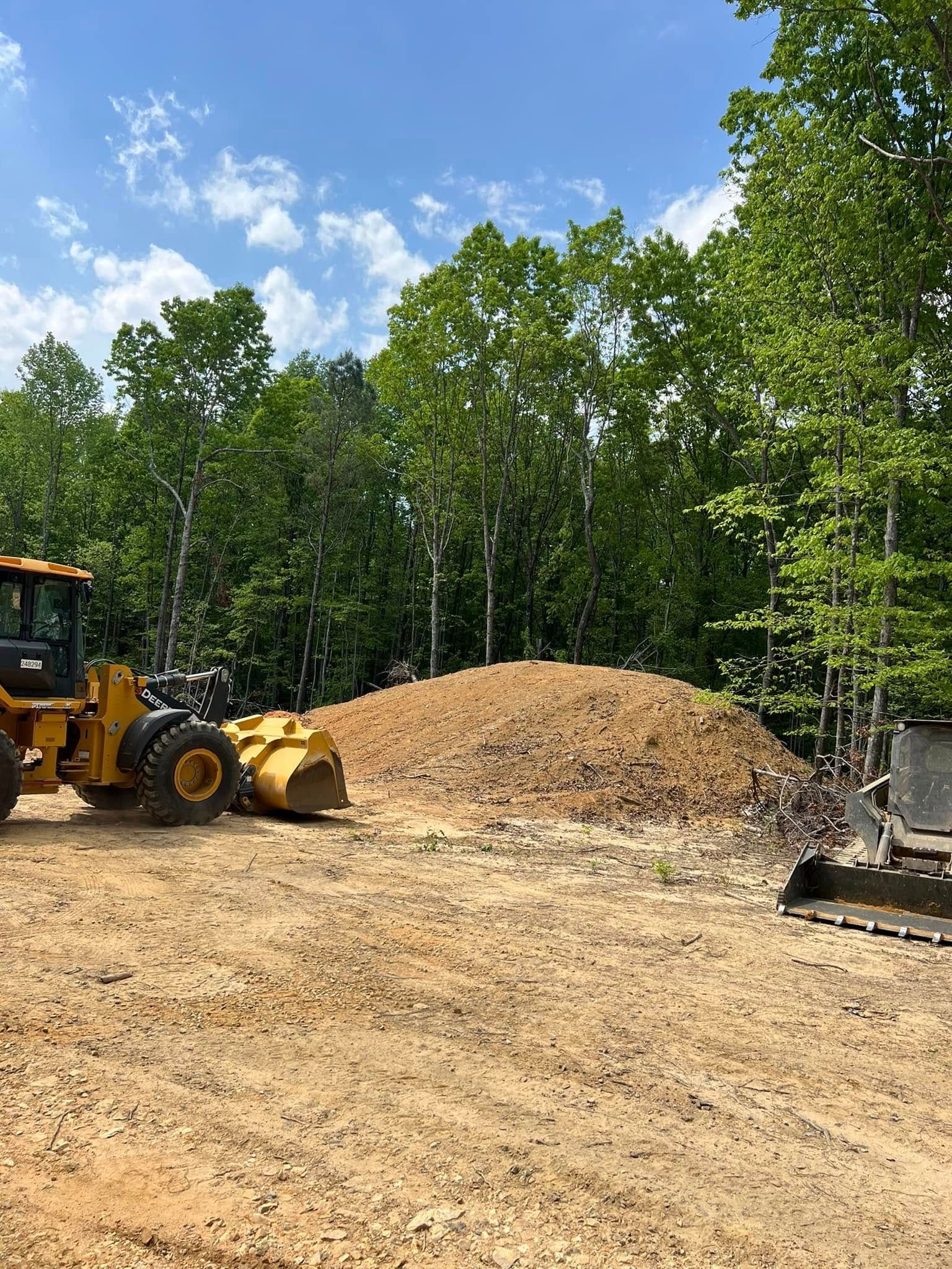Yellow construction vehicle parked by a large dirt mound in a wooded area under a blue sky.