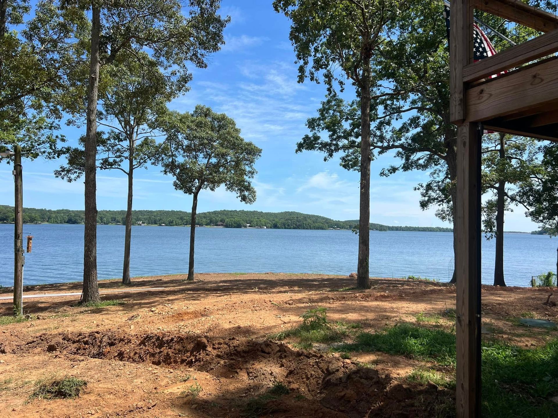 A lake view from a backyard with freshly cleared soil, towering trees, and a wooden deck corner under a blue sky.