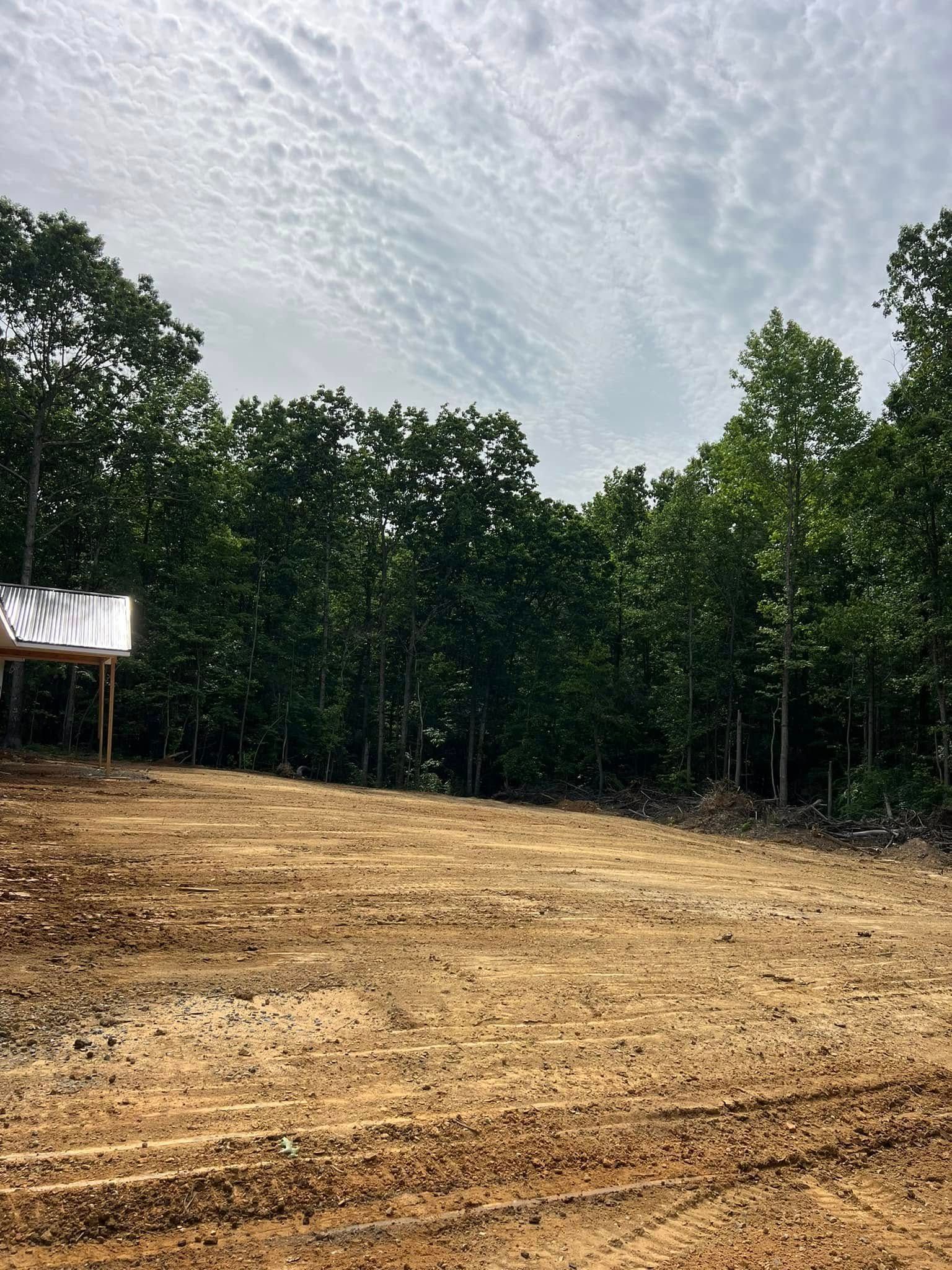 A cleared, dirt construction site borders a dense green forest under a cloudy sky.