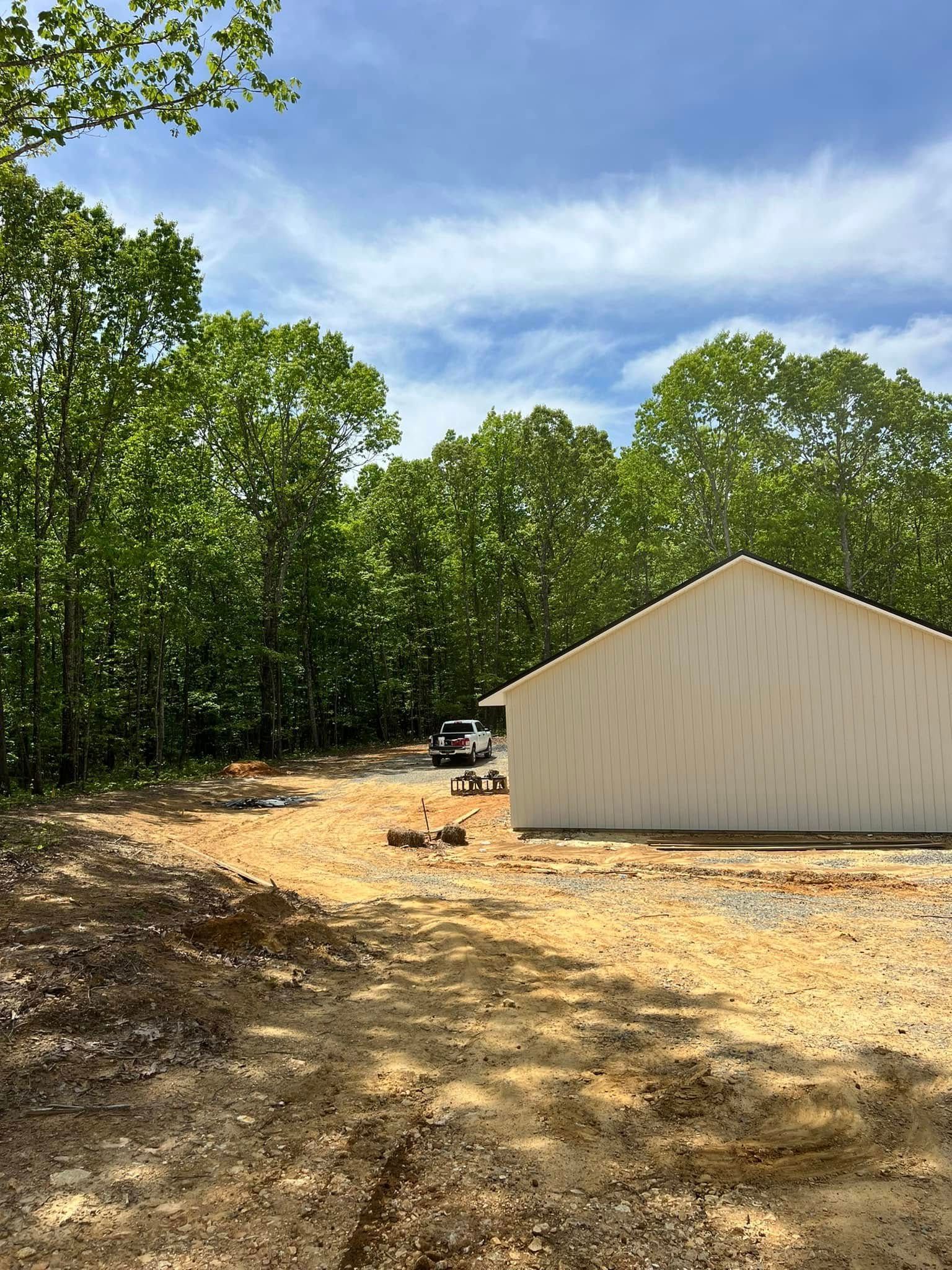 A white metal building sits on a gravel lot bordered by a dense forest under a bright blue sky.