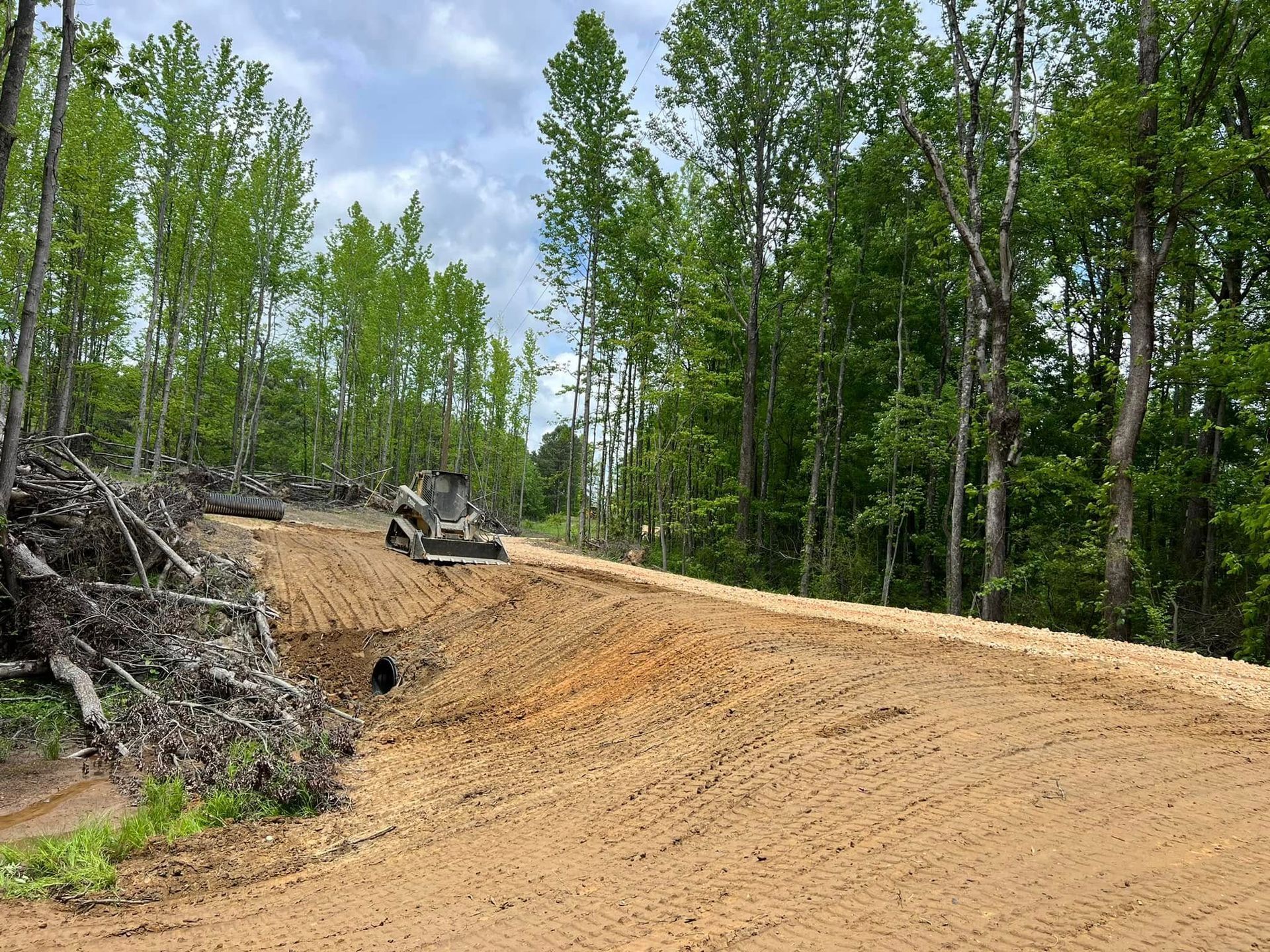 A small bulldozer works on a graded dirt road through a forested area with a pile of cleared branches to the left.