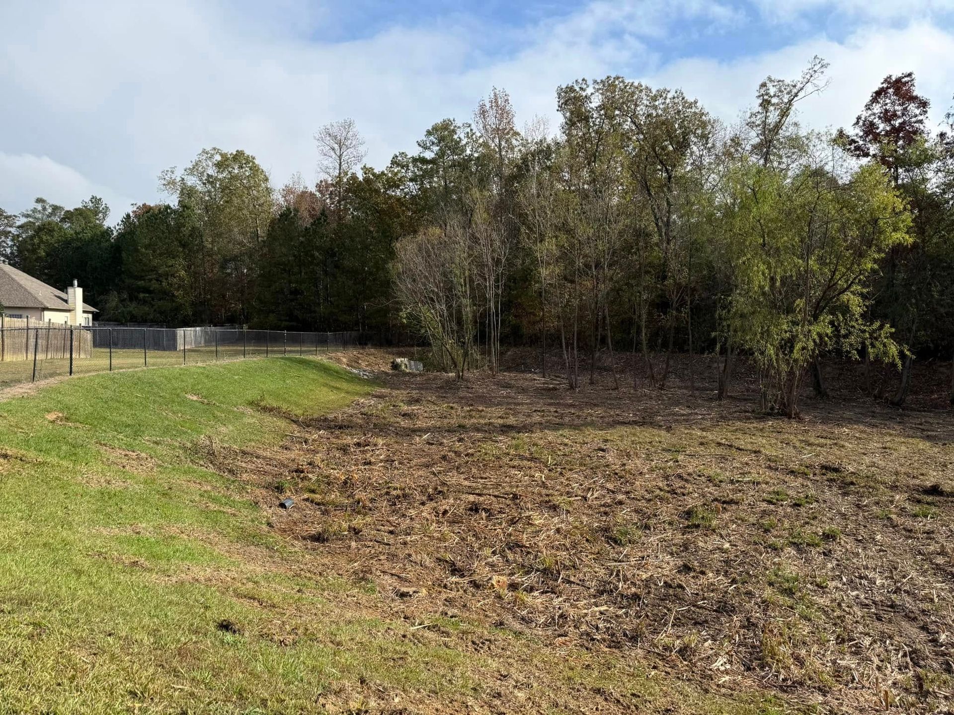 A cleared, brown dirt lot slopes upward toward a dense treeline, with a grassy berm and a house fence on the left.