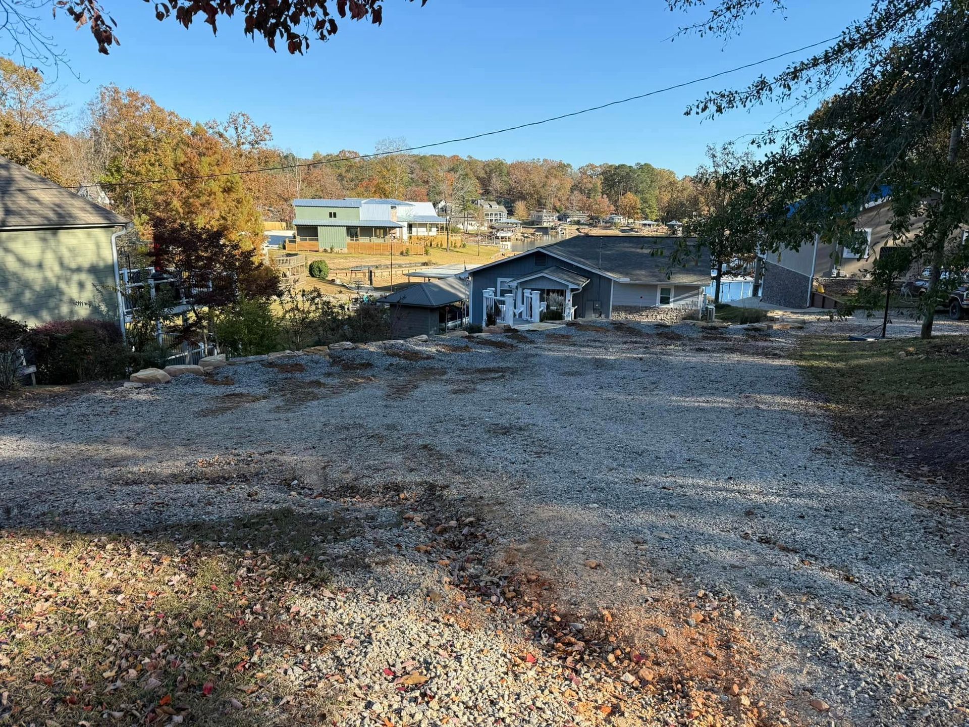 A gravel driveway slopes downward toward suburban houses on a sunny day with trees in autumn foliage.