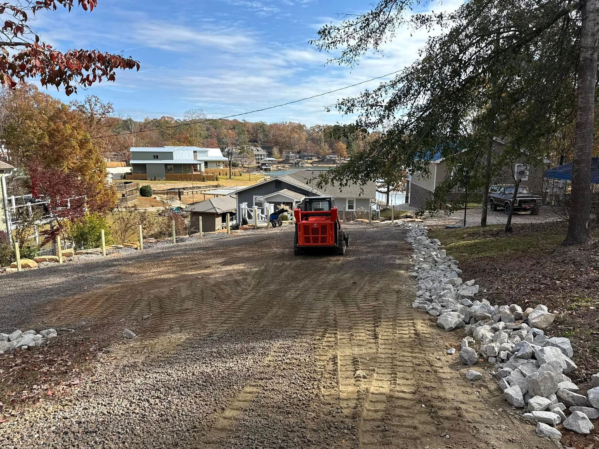 A red skid steer loader sits on a gravel driveway in a residential neighborhood under a blue sky.