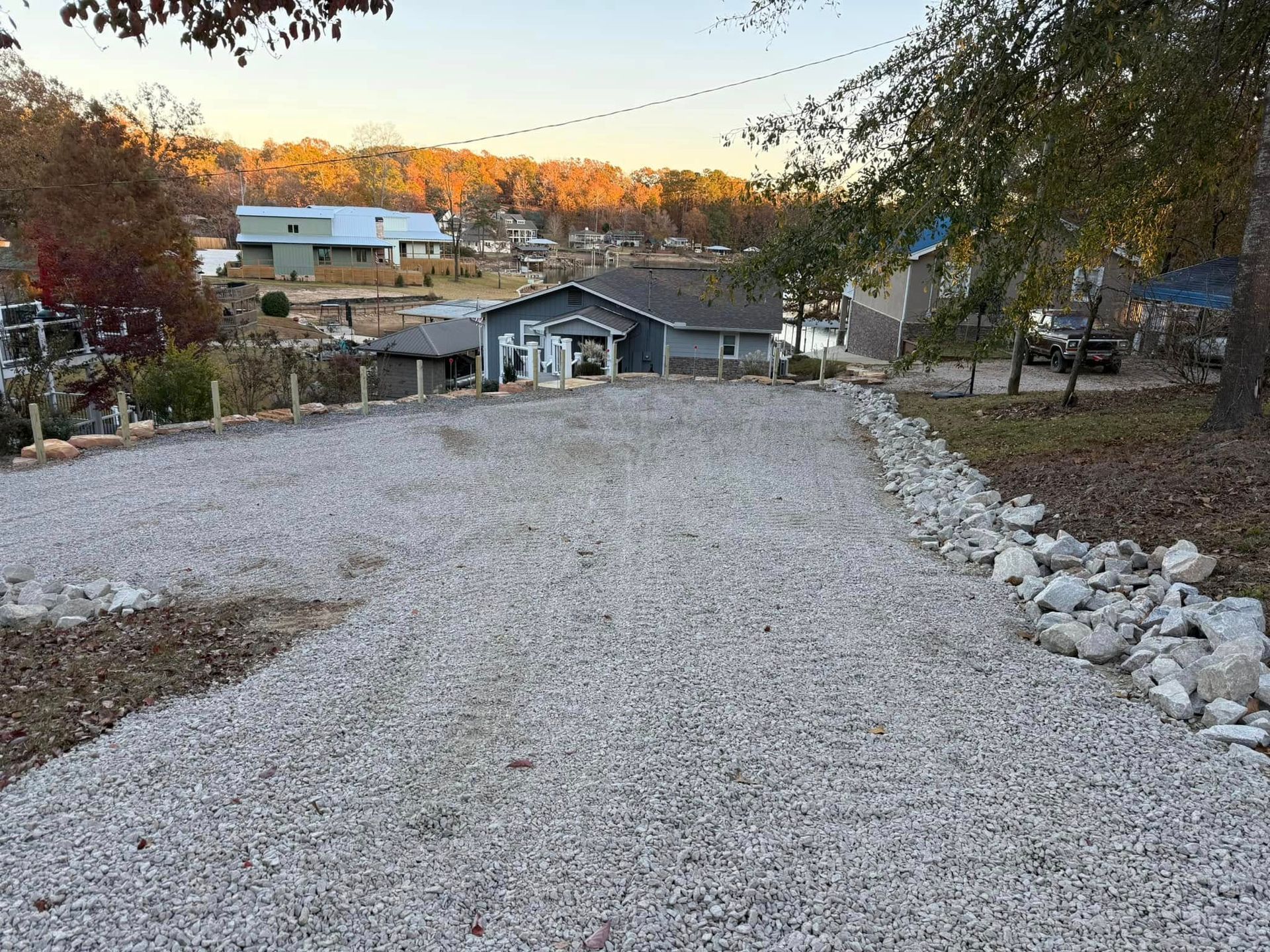A gravel driveway leads toward a house at sunset, bordered by trees and a line of rocks on the right side.