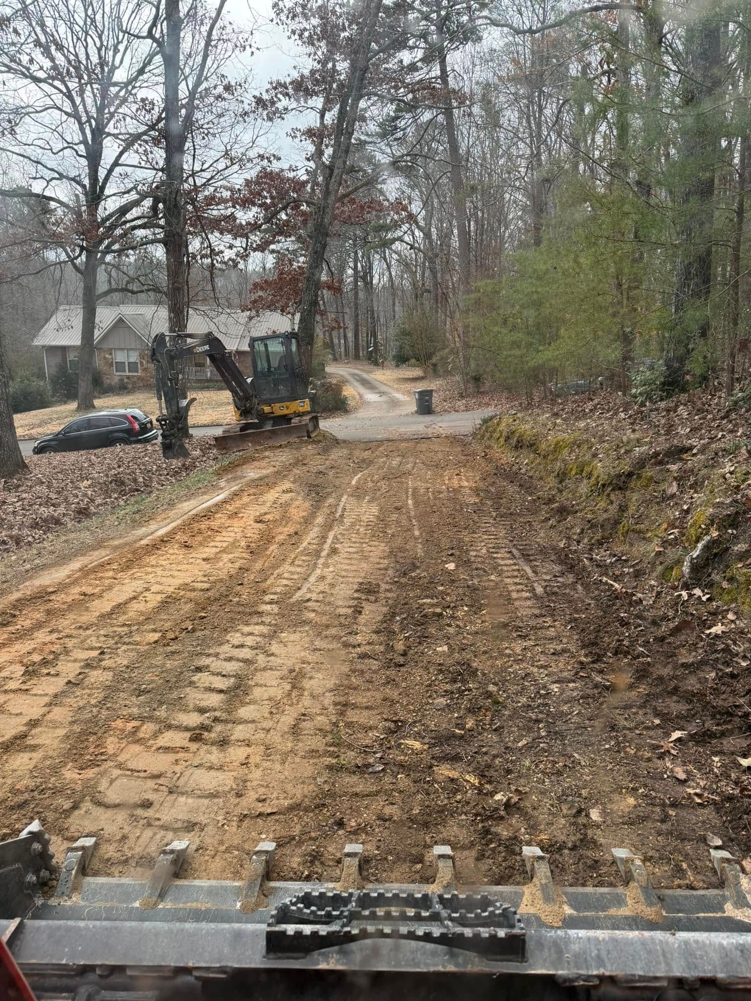 An excavator viewed from the cab, overlooking a dirt path leading toward a house and wooded area.