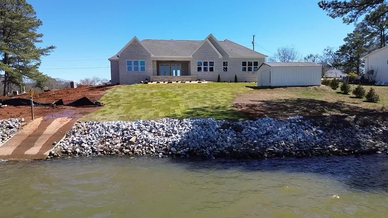 A beige house sits above a rock-lined waterfront with a boat ramp and grass lawn on a sunny day.