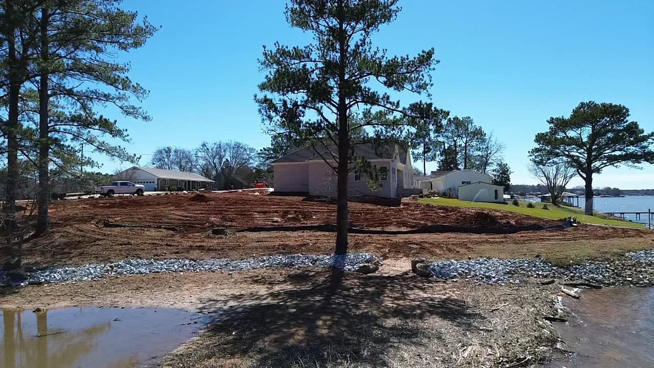 A waterfront property under construction with a cleared red-dirt yard, pine trees, and a house frame near the shoreline.