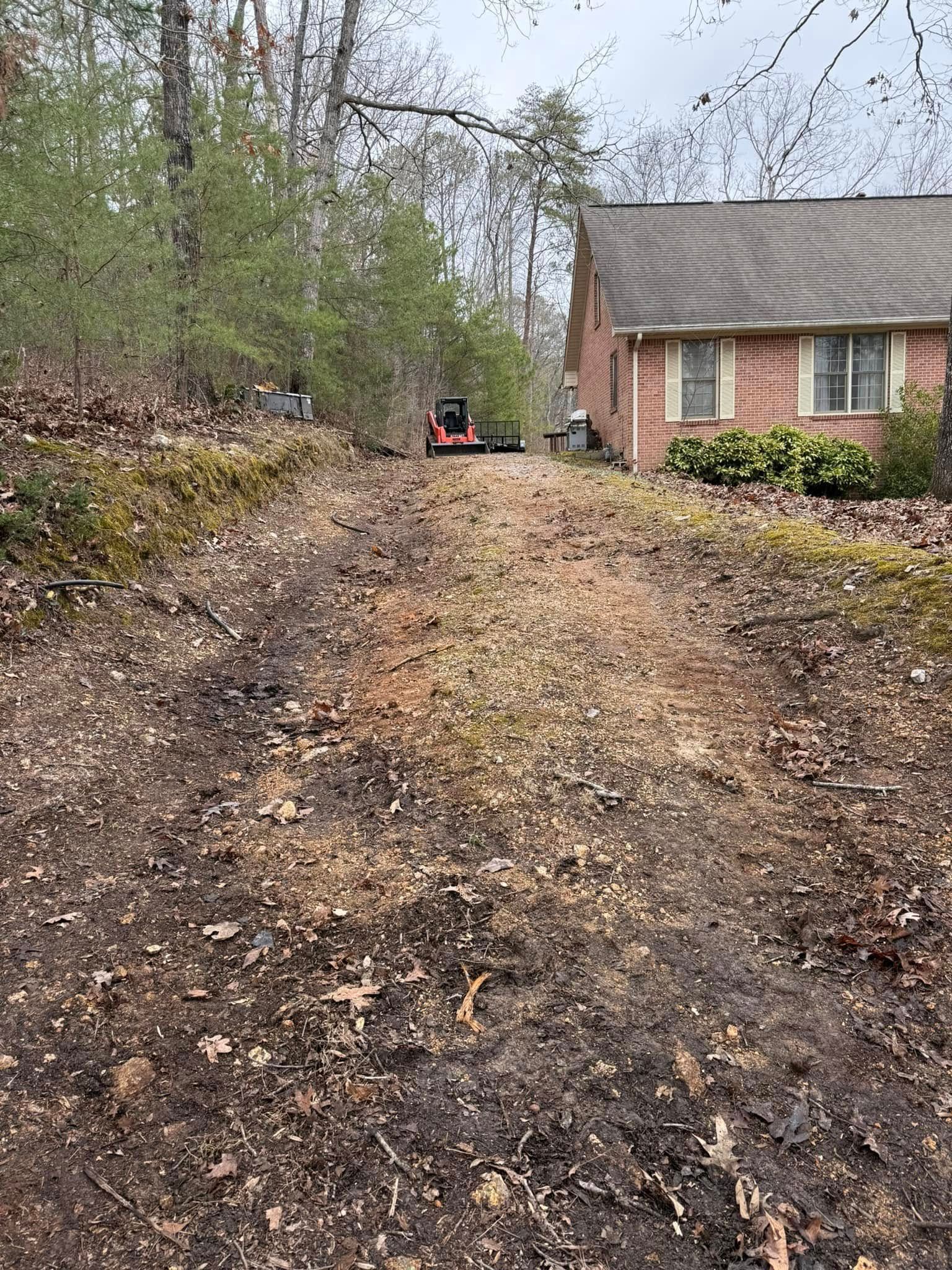 A dirt path leads through a wooded area toward the side of a brick house with a red tractor visible in the distance.