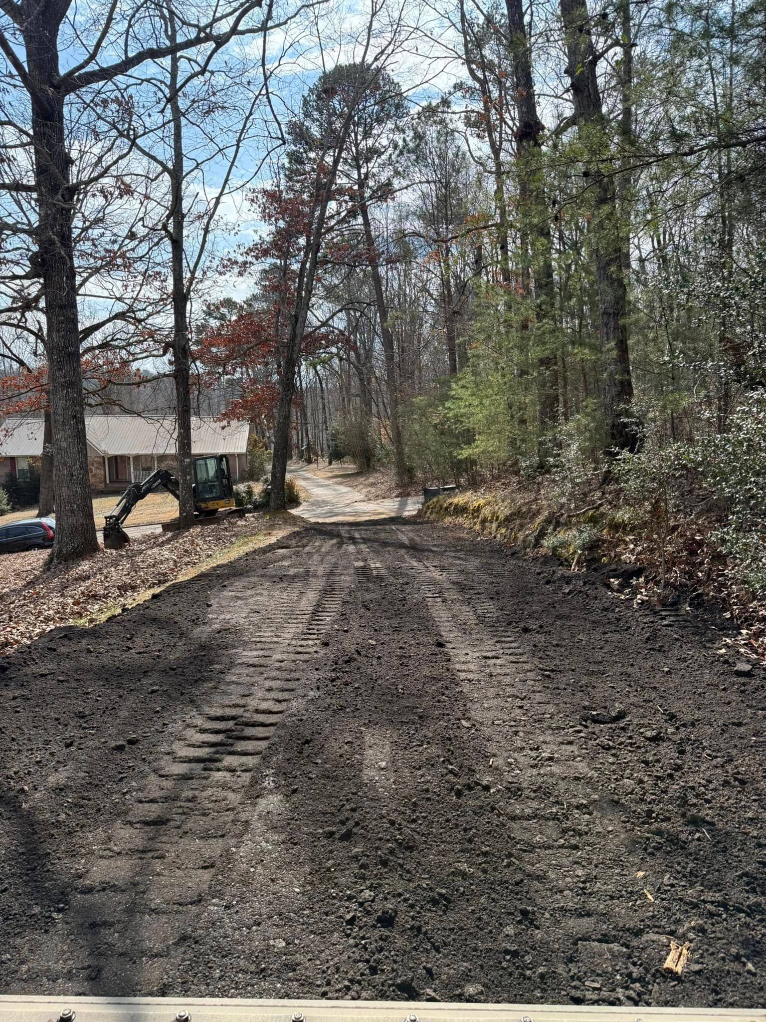 A construction excavator sits at the edge of a cleared, muddy dirt path leading into a wooded residential area.