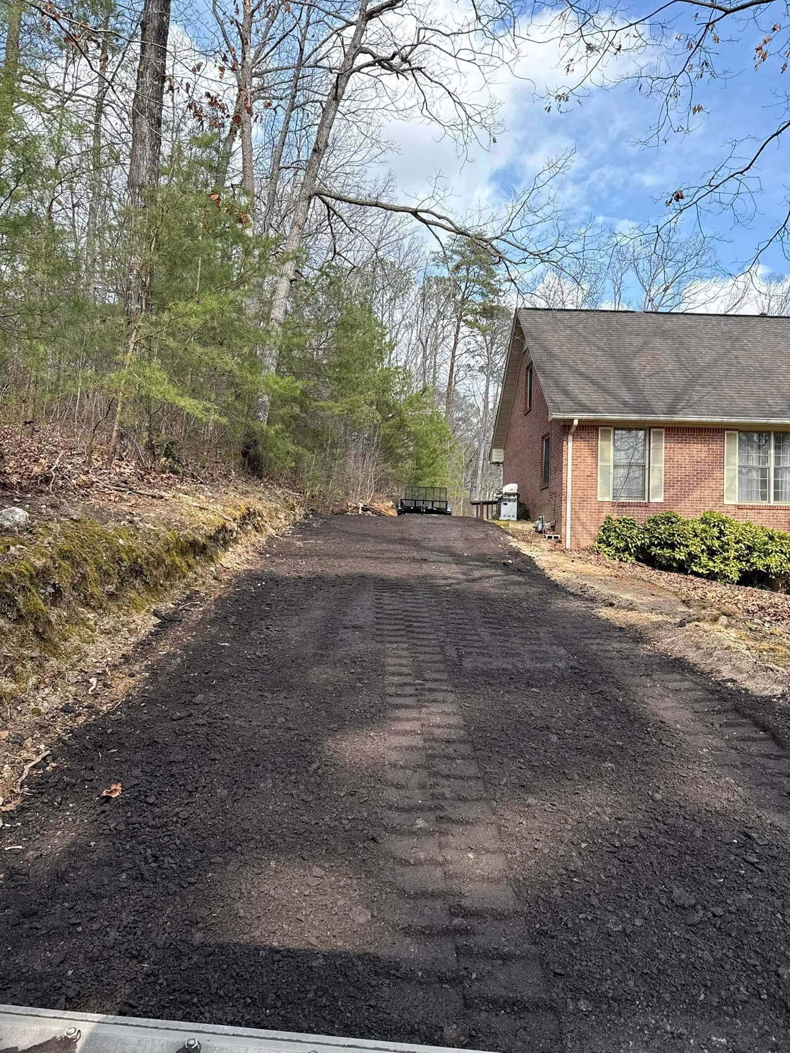 A newly paved gravel driveway leads toward the side of a brick house next to a wooded area.