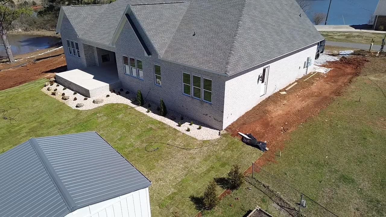An aerial view of a gray-roofed house with a white exterior, featuring a stone patio and surrounding yard.