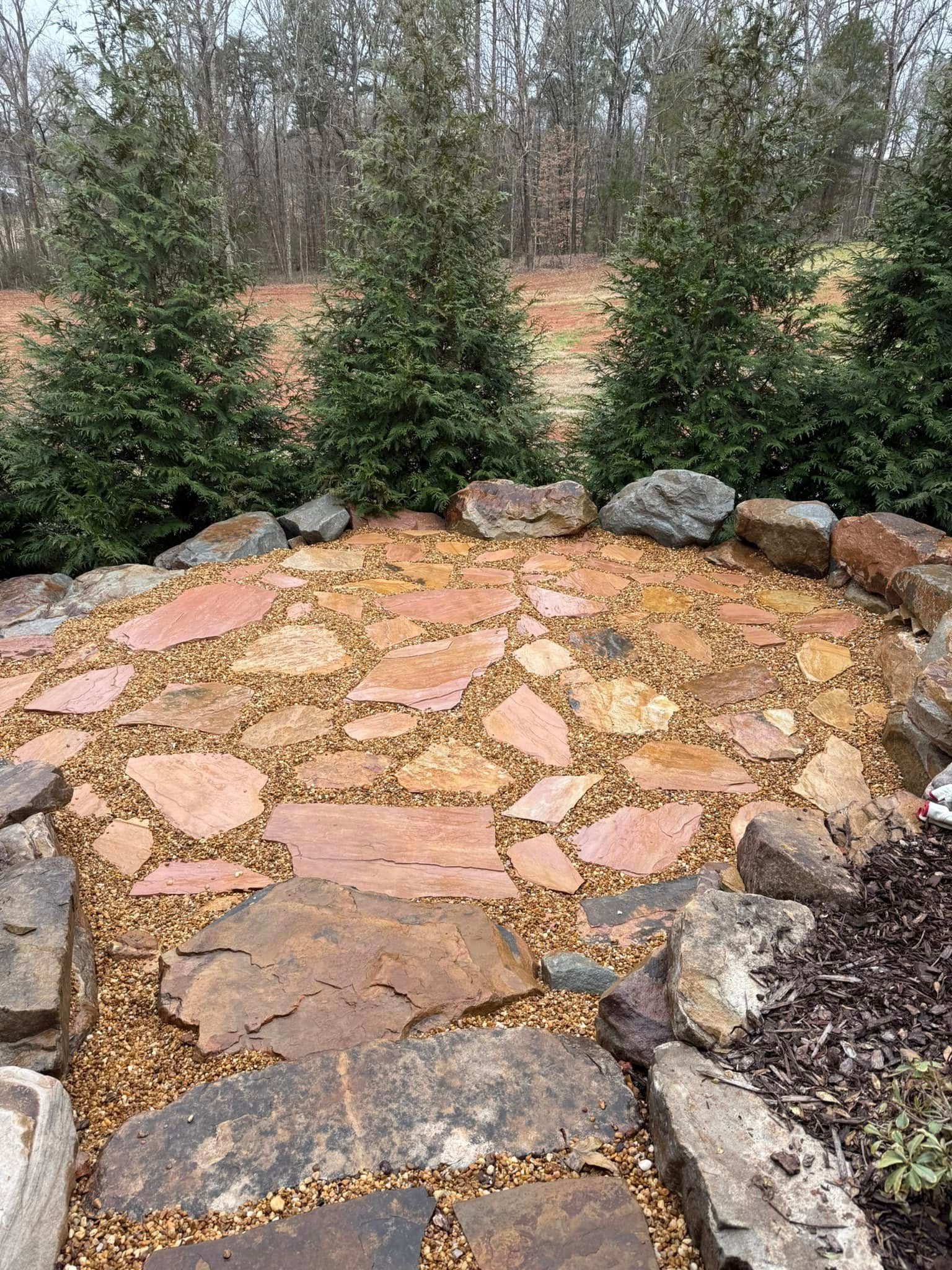 A stone patio made of irregular tan flagstones set in gravel, bordered by large rocks and three evergreen trees.