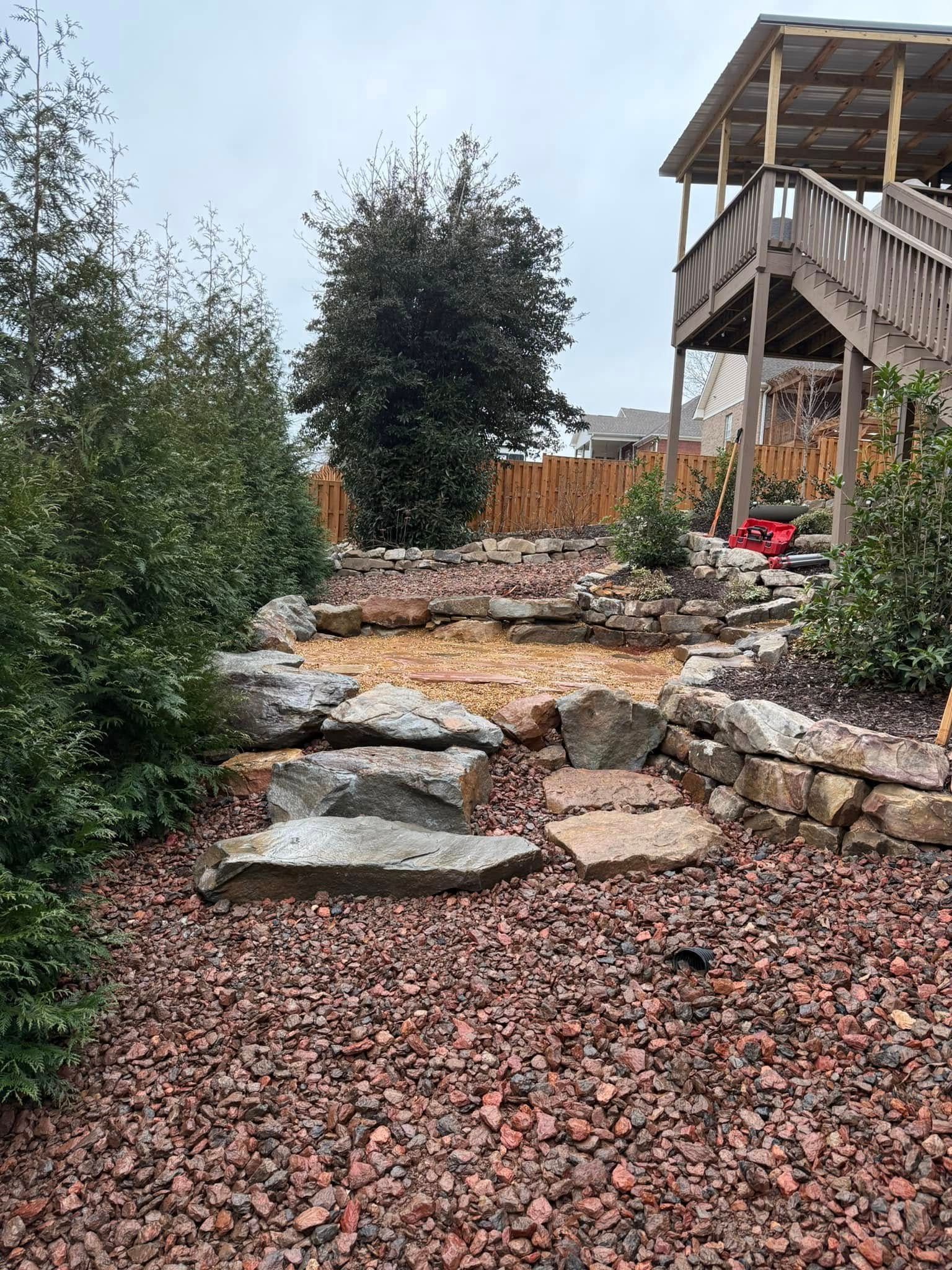 A landscape bed featuring green shrubs and small plants growing in bright red mulch next to a tree trunk.