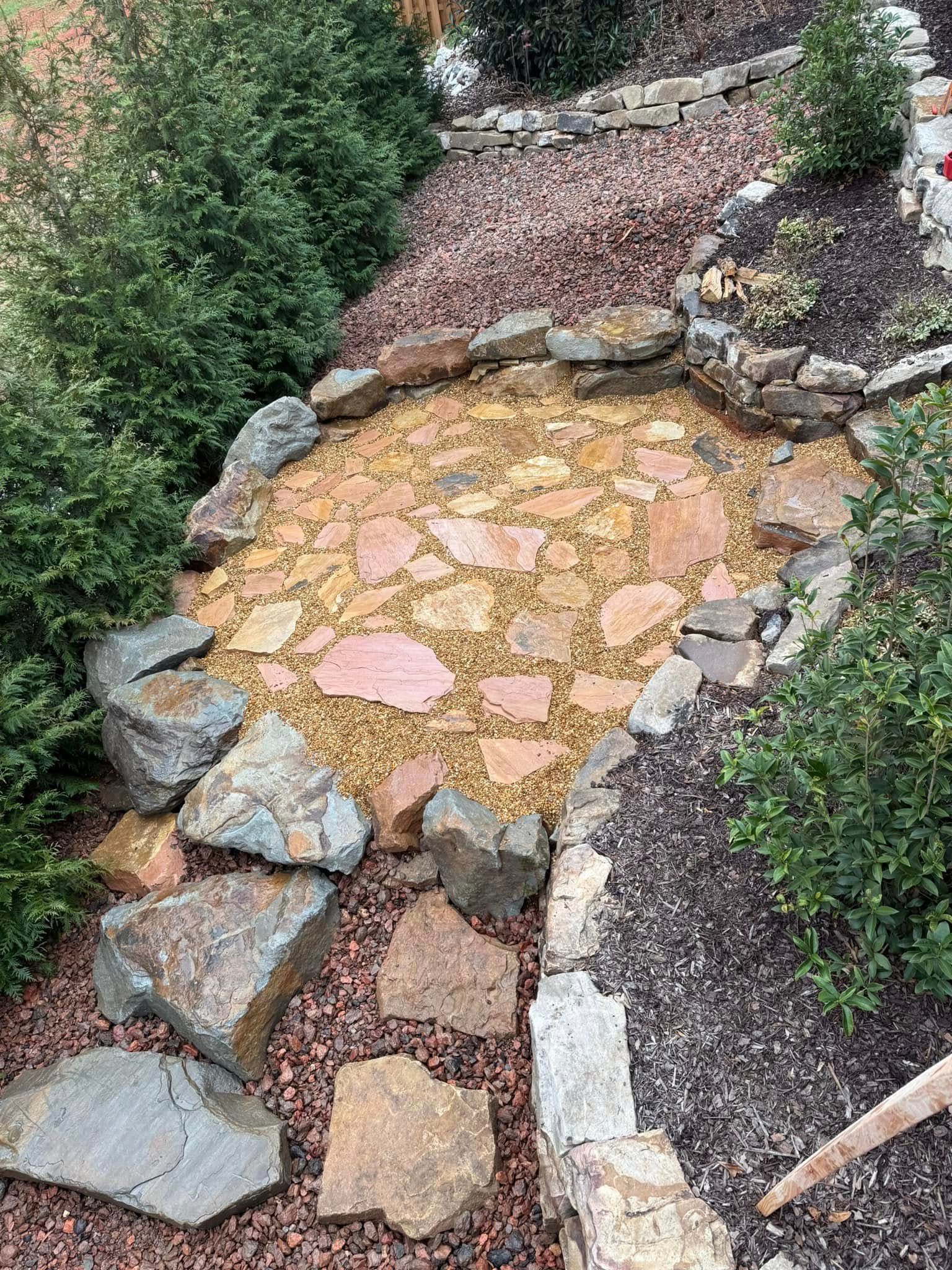 A stone-bordered garden path with a circular area of flagstones set in tan gravel, surrounded by shrubs and dark mulch.