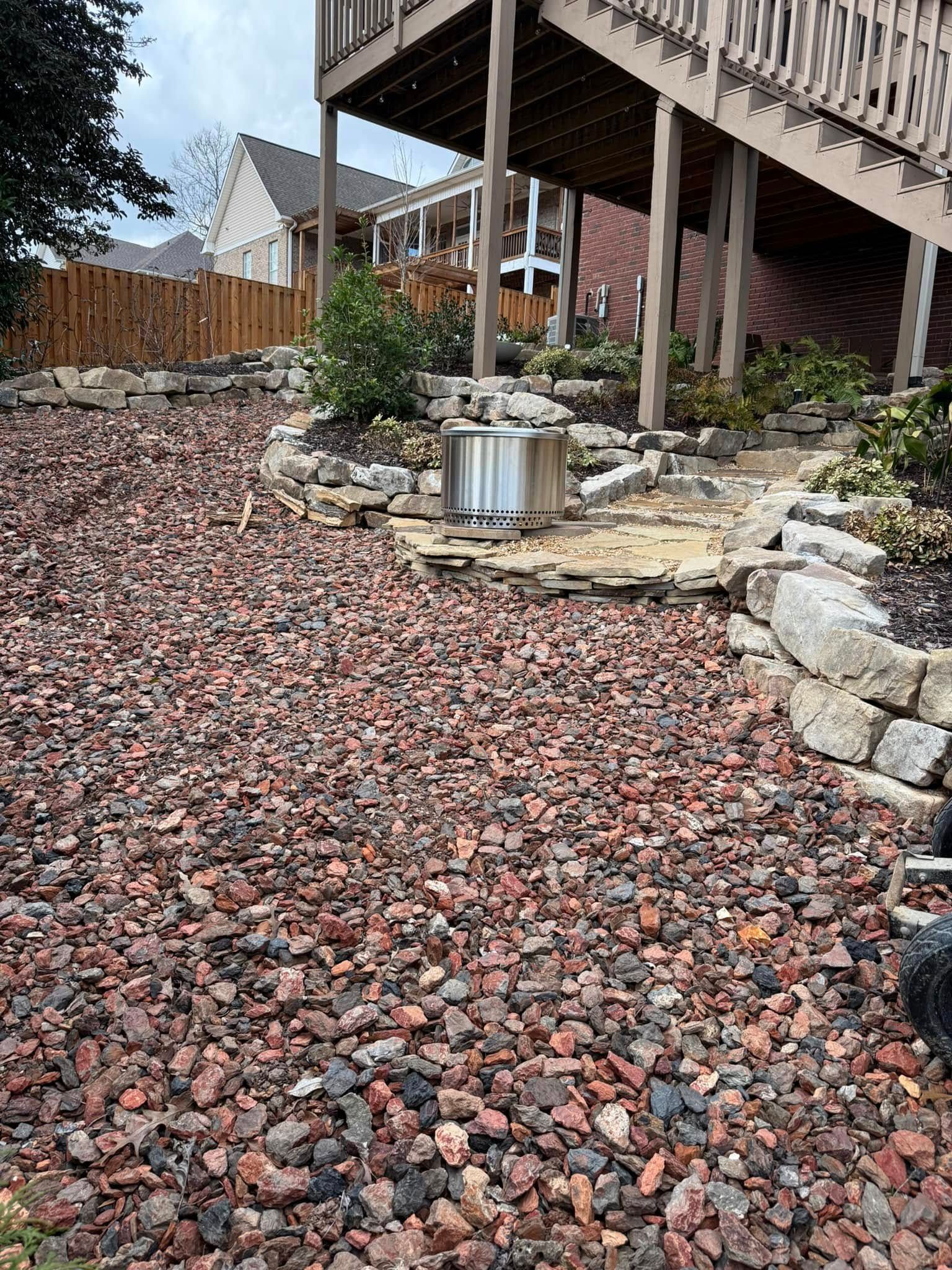 A metal fire pit sits on a stone patio beneath a wooden deck, surrounded by a yard covered in reddish-brown gravel.
