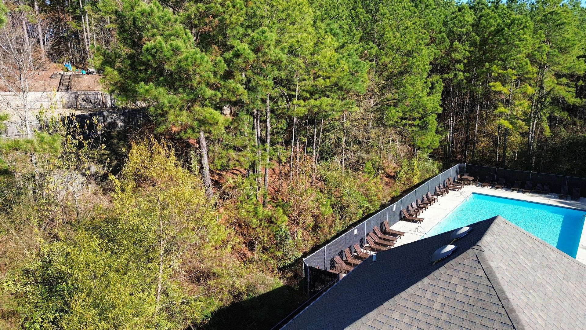 An aerial view shows a swimming pool with a row of lounge chairs next to a building roof and dense, green woodland.