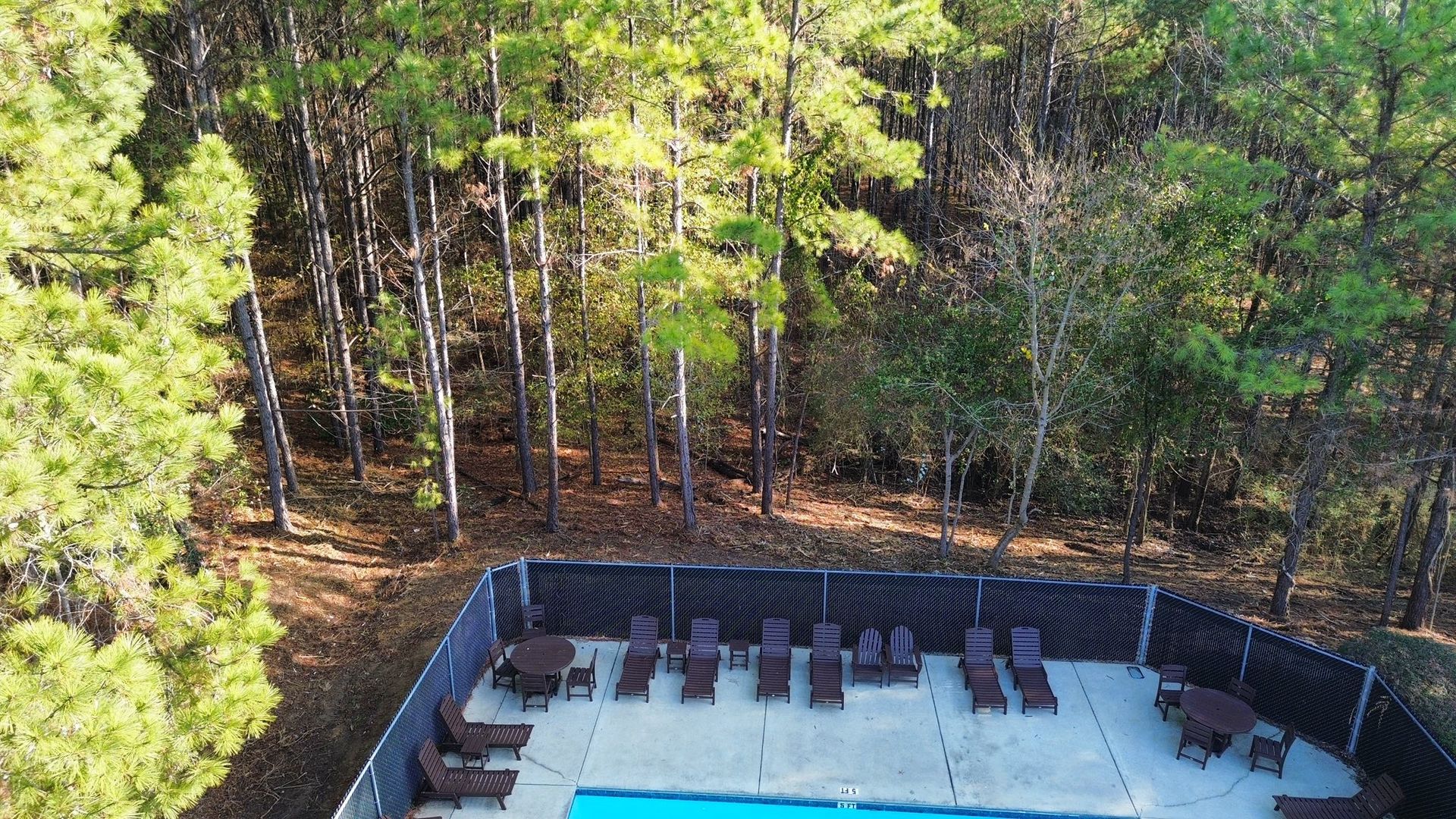 An elevated view of a swimming pool deck lined with lounge chairs, bordered by a dark privacy fence and a dense forest.