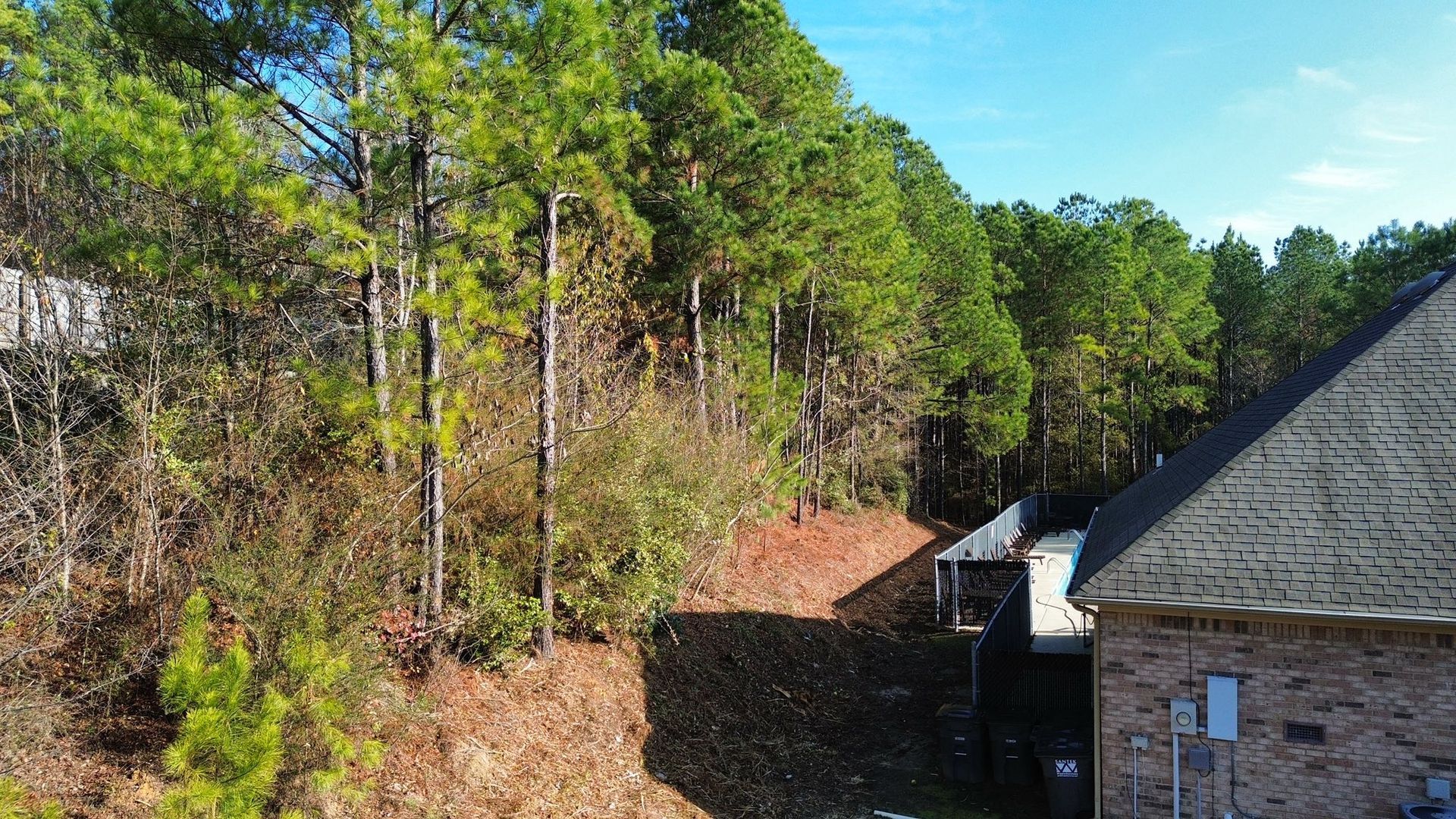 A view of a house exterior beside a steep, wooded hillside covered in dry leaves under a clear blue sky.