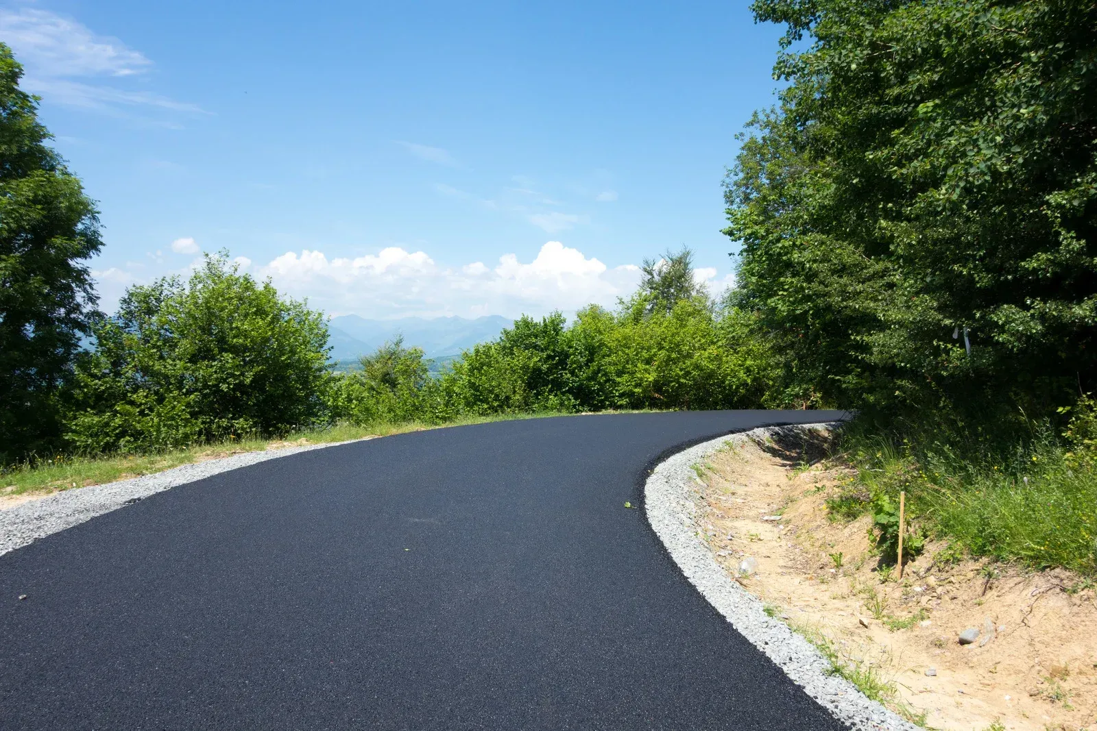 A fresh, black asphalt road curves through a sunny, lush green landscape with distant mountains under a blue sky.