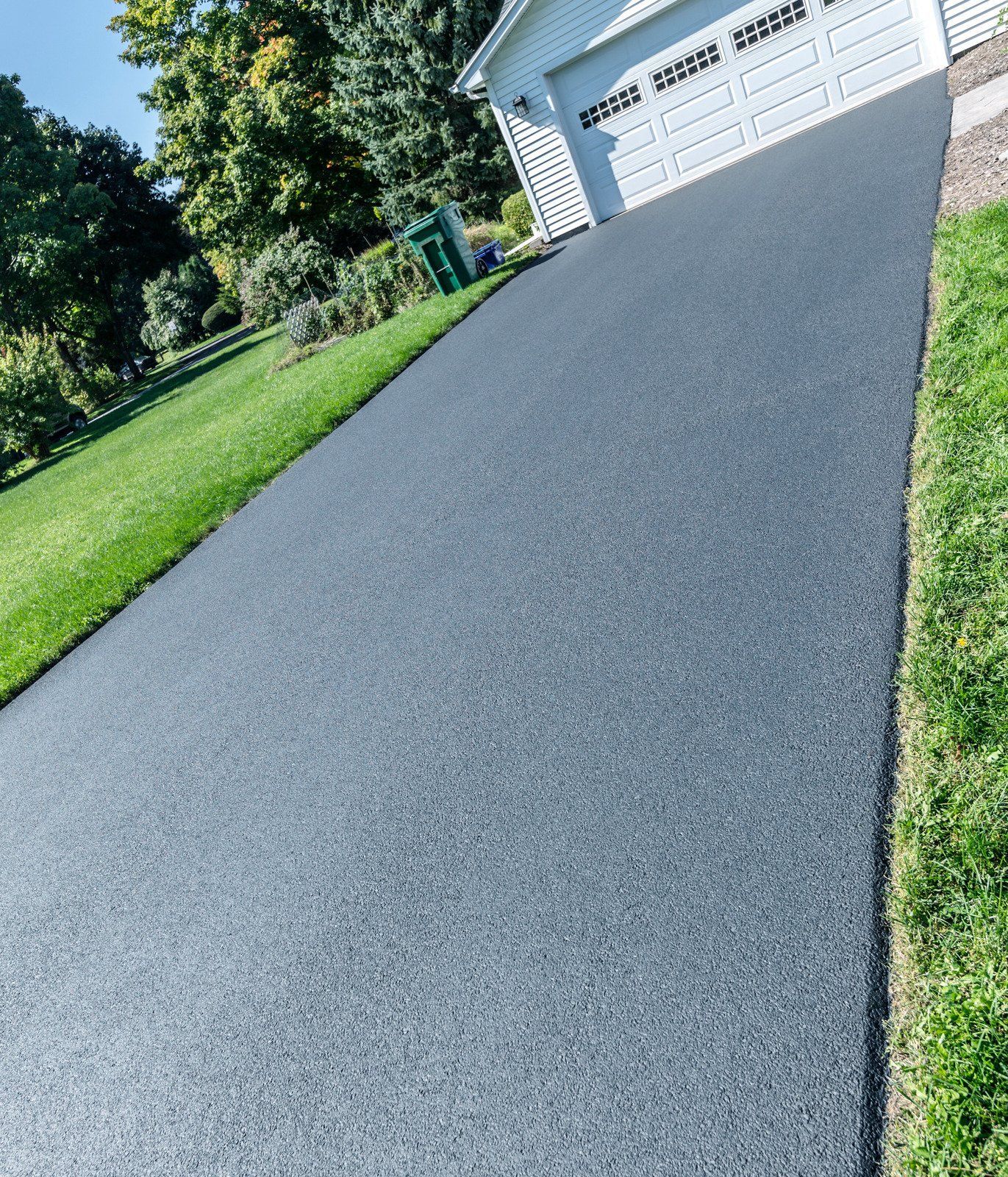 A freshly paved, dark gray asphalt driveway leading up to a white garage door, framed by green grass and trees.