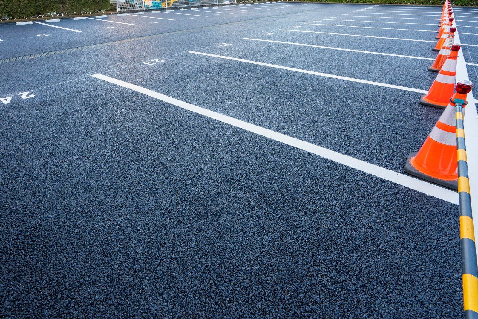 A row of orange and white safety cones connected by a tape barrier lines the side of an empty asphalt parking lot.