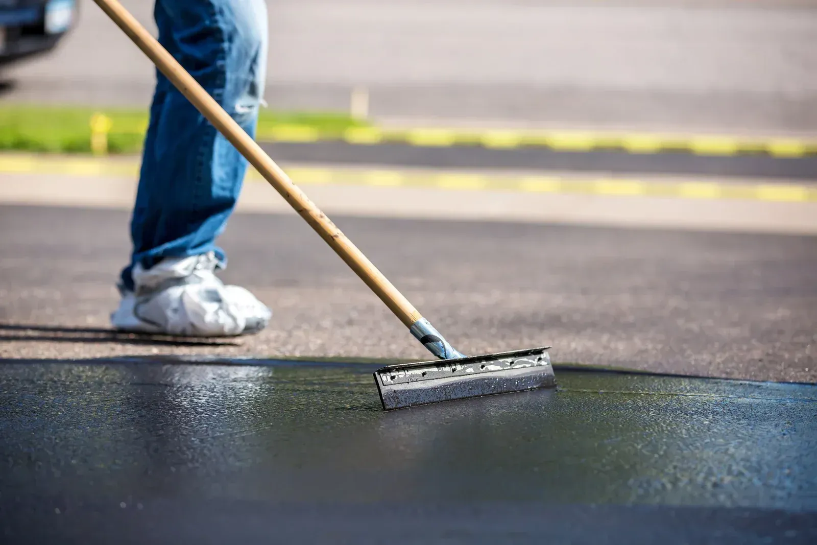 A person in jeans uses a squeegee to spread dark, liquid asphalt sealant evenly across a paved driveway.