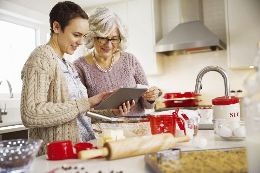 A home health aide assists an elderly woman cooking using an tablet.