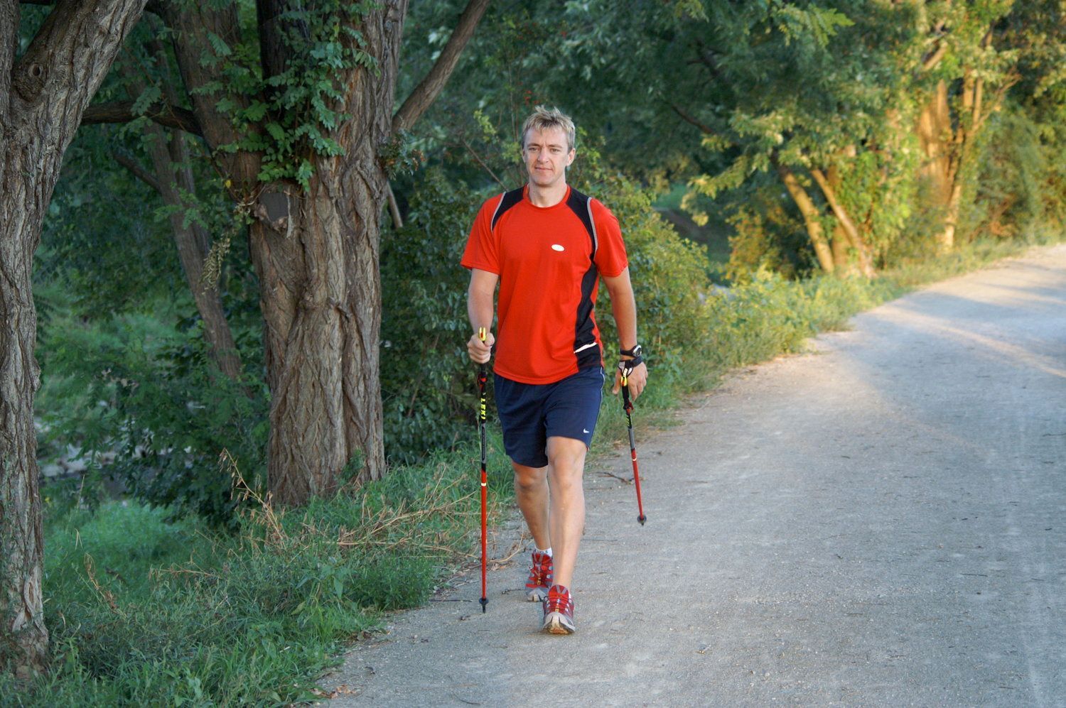 A man in a red shirt is walking down a road with walking poles.