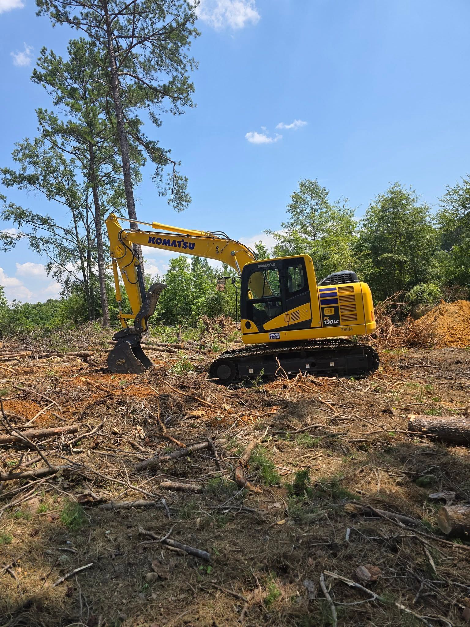 Yellow Komatsu excavator clearing land, against a blue sky, amidst tree debris.