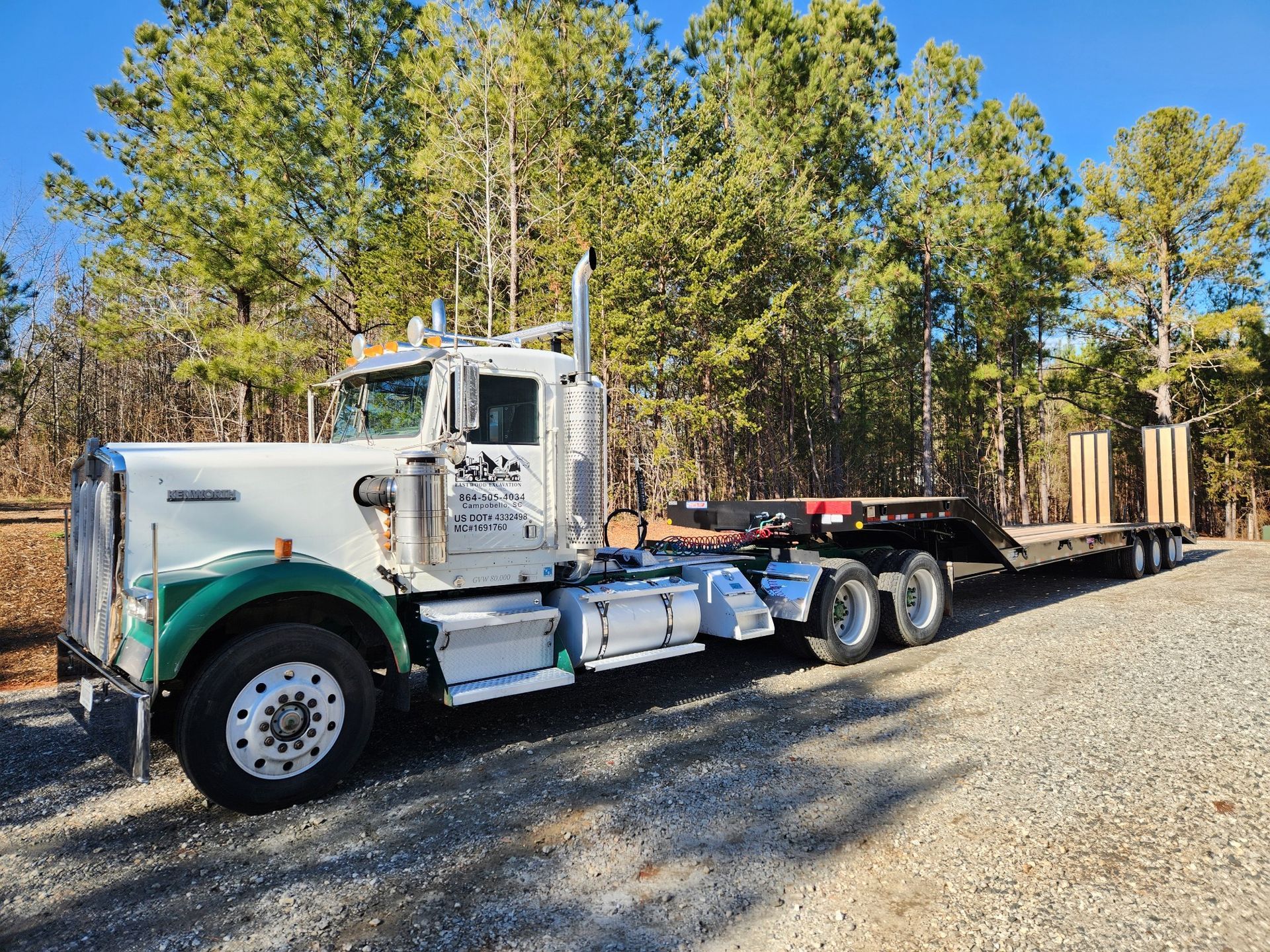 White and green semi-truck with flatbed trailer hauling lumber, parked on gravel.