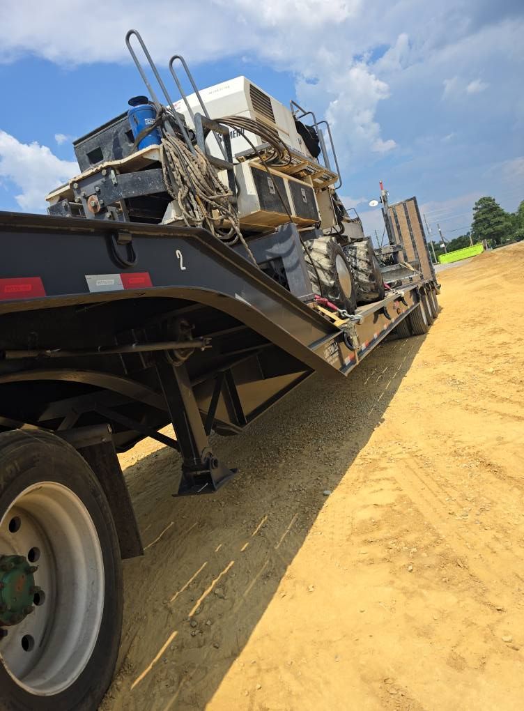 Flatbed trailer hauling heavy construction equipment on a dirt lot.