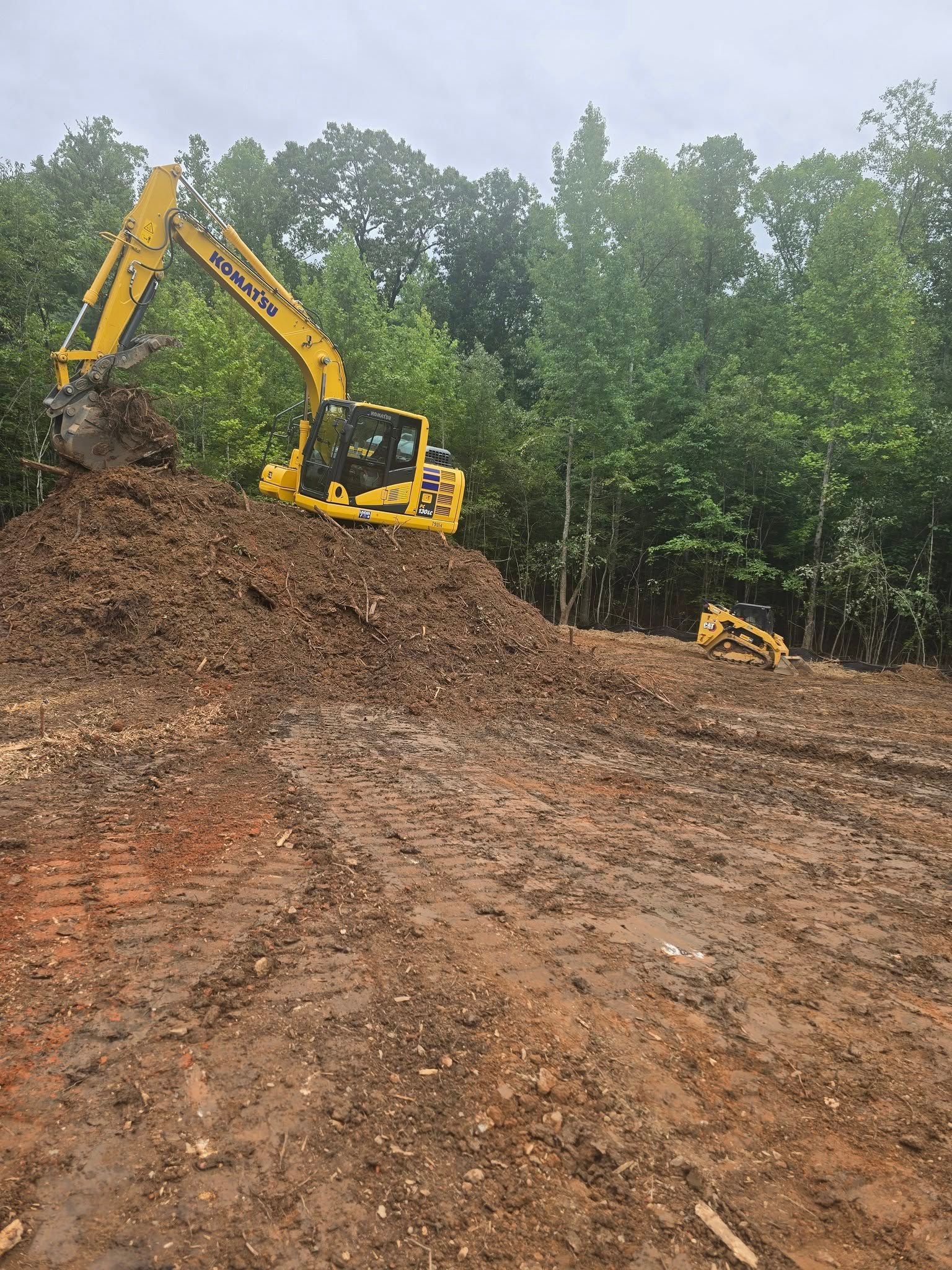 Yellow excavator on a muddy site, digging dirt. Trees in background.