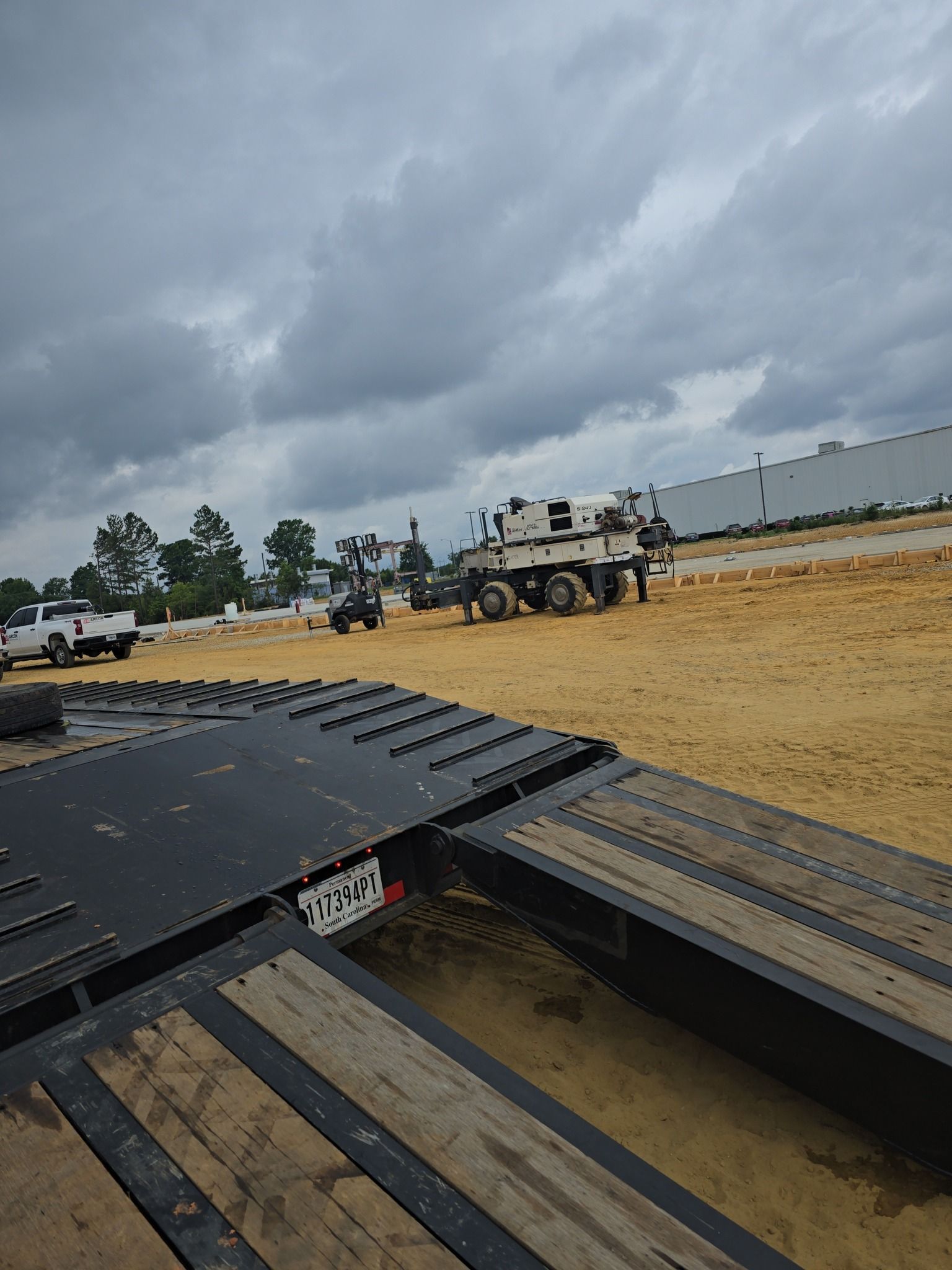 A flatbed trailer with a crane in the background on a dirt lot under a cloudy sky.