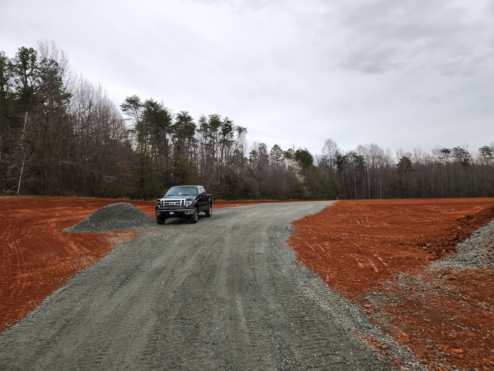 Black truck on a gravel road in a cleared area with red soil, surrounded by trees under a cloudy sky.