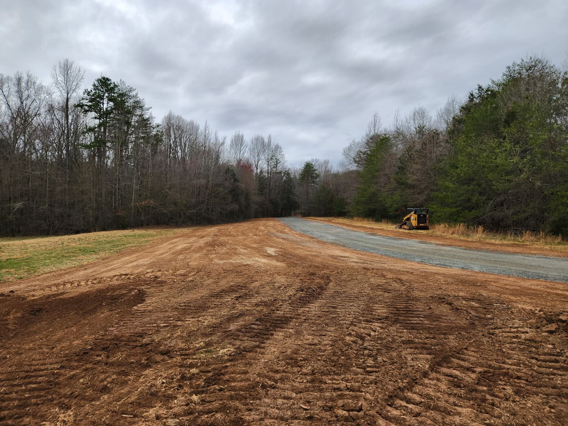 Dirt field with freshly graded dirt path, gravel road, and trees under cloudy sky.